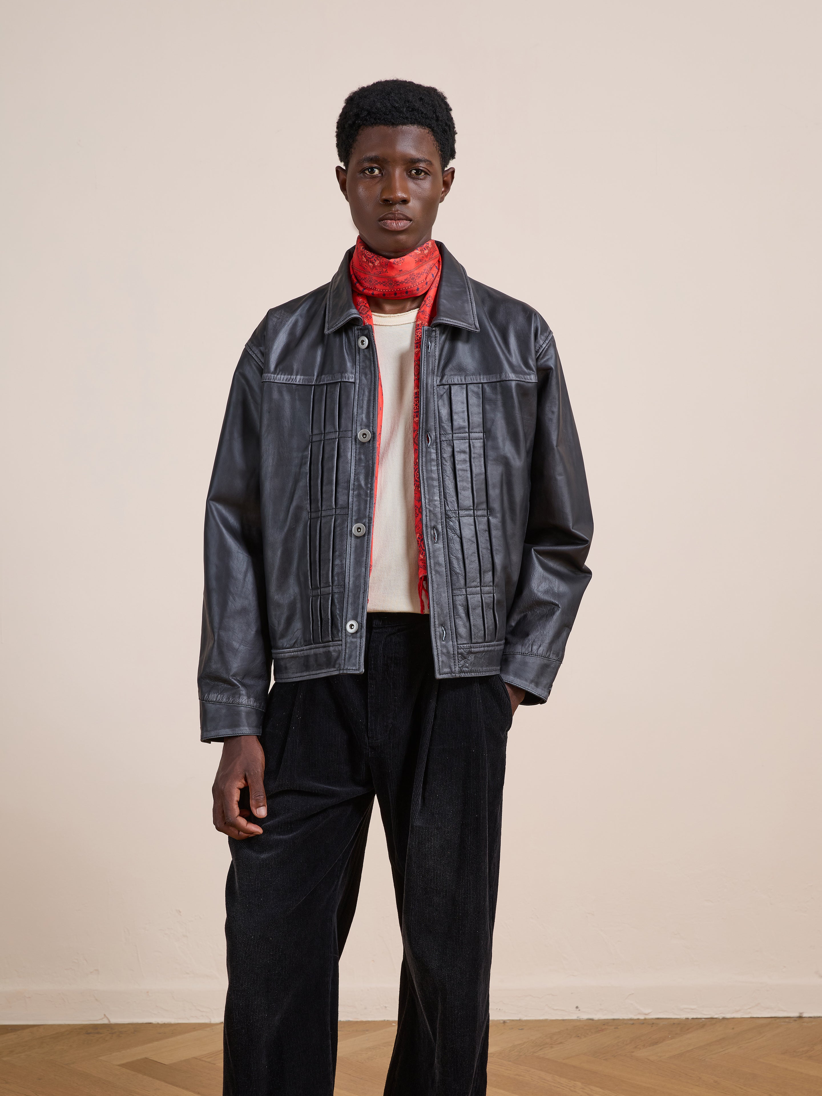 Indoors, a person wears the FOUND Trucker Leather Jacket with a white shirt, black pants, and a red scarf, standing against a plain light wall.