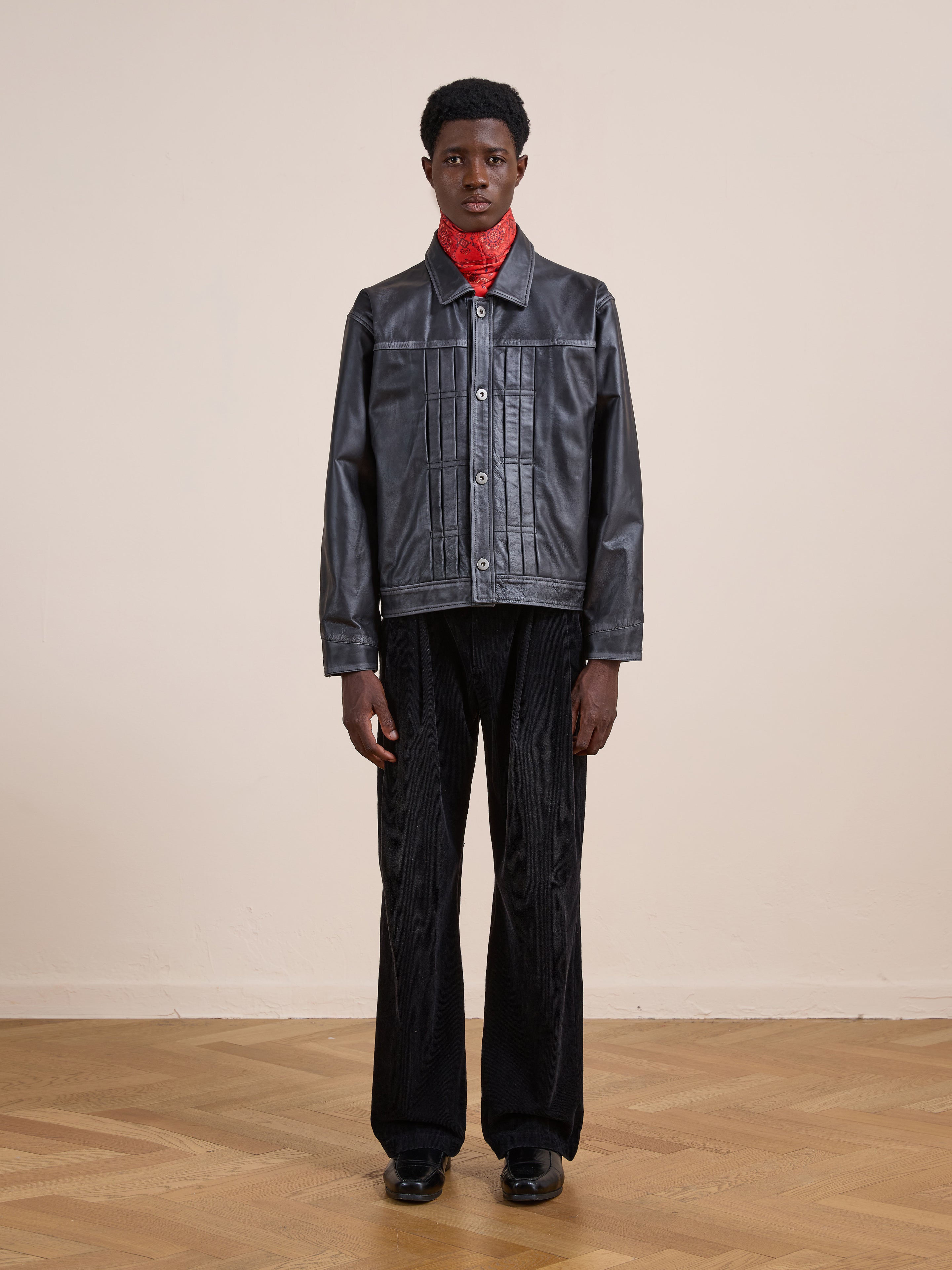 Indoors on a wooden floor, a person wears the FOUND Trucker Leather Jacket in vintage black with a relaxed fit, styled with black pants, black shoes, and a red bandana, standing against a plain light-colored wall.