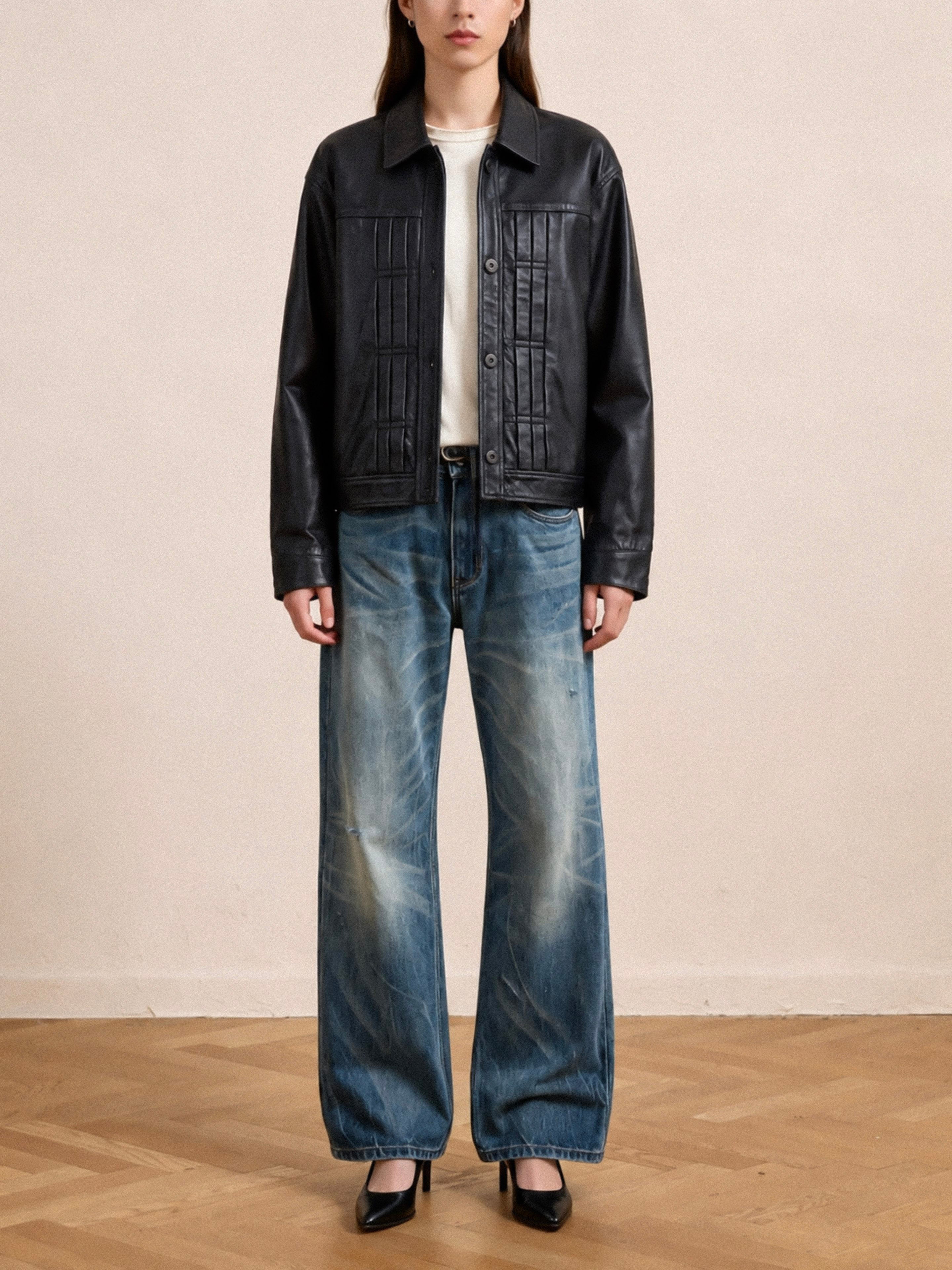 A person stands indoors on a wooden floor in front of a plain beige wall, wearing the FOUND Trucker Leather Jacket styled with a white shirt, wide-leg blue jeans, and black heels.