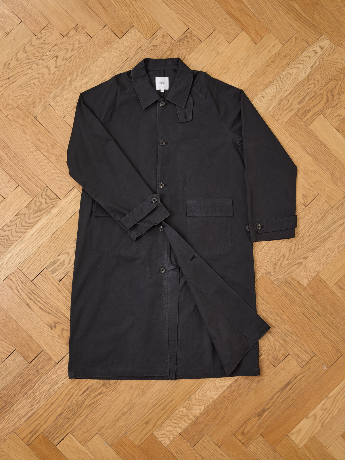 A FOUND Twill Work Trench Coat in black cotton twill, featuring a belt, is laid flat on a light wood herringbone floor.