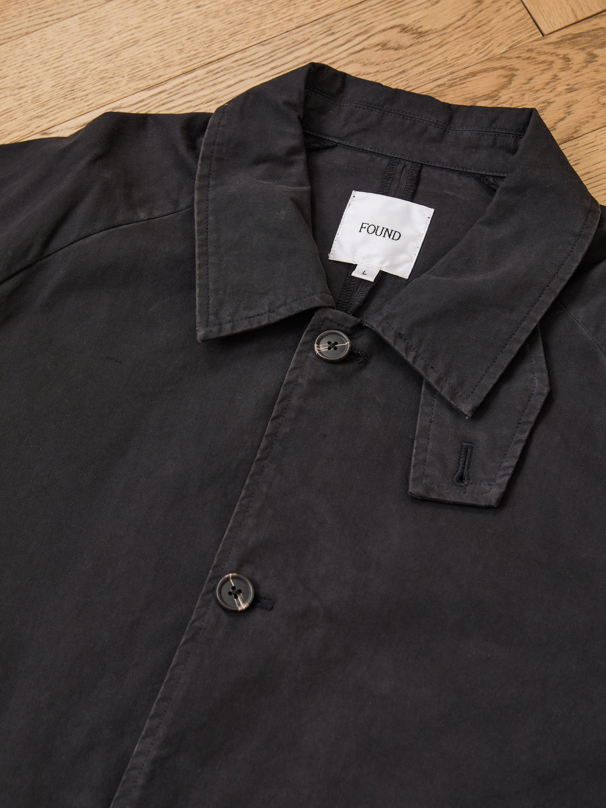 FOUND Twill Work Trench Coat—a black unisex button-up, collared jacket laid flat on a wooden floor, designed for everyday versatility.