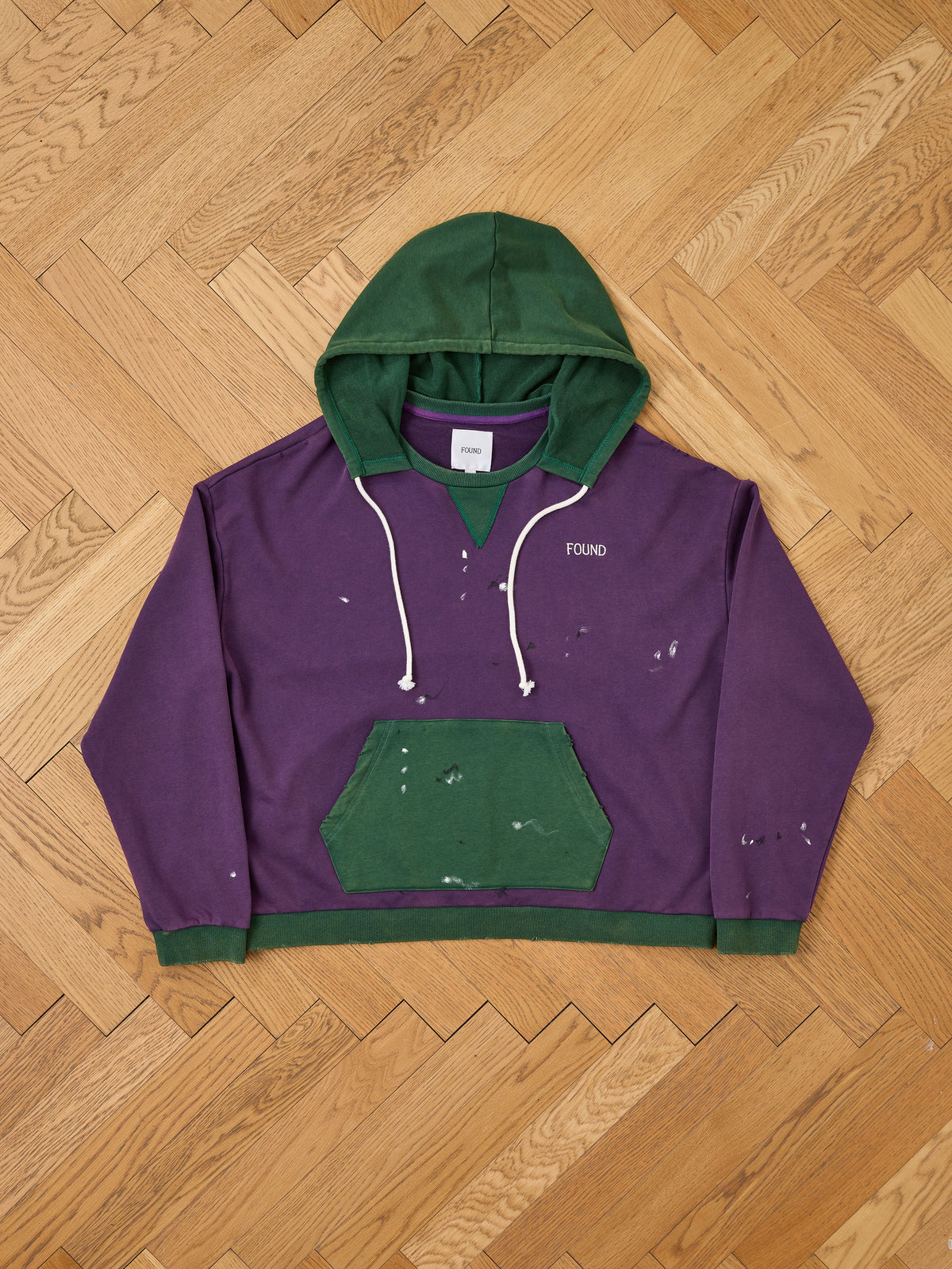 The Two Tone Painter Hoodie by FOUND features purple and green distressed cotton, white drawstrings, “FOUND” text, paint splatters, and a kangaroo pocket on herringbone wood.