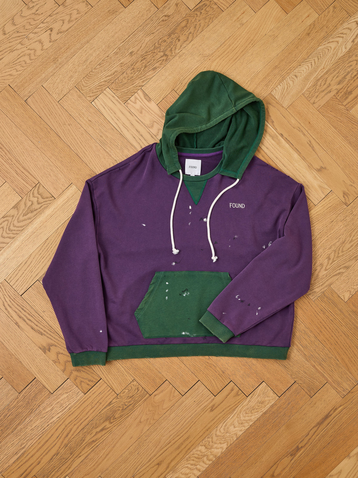 The FOUND Two Tone Painter Hoodie in purple and green with white paint splatters features a relaxed unisex fit, “FOUND” on the chest, and a kangaroo pocket.