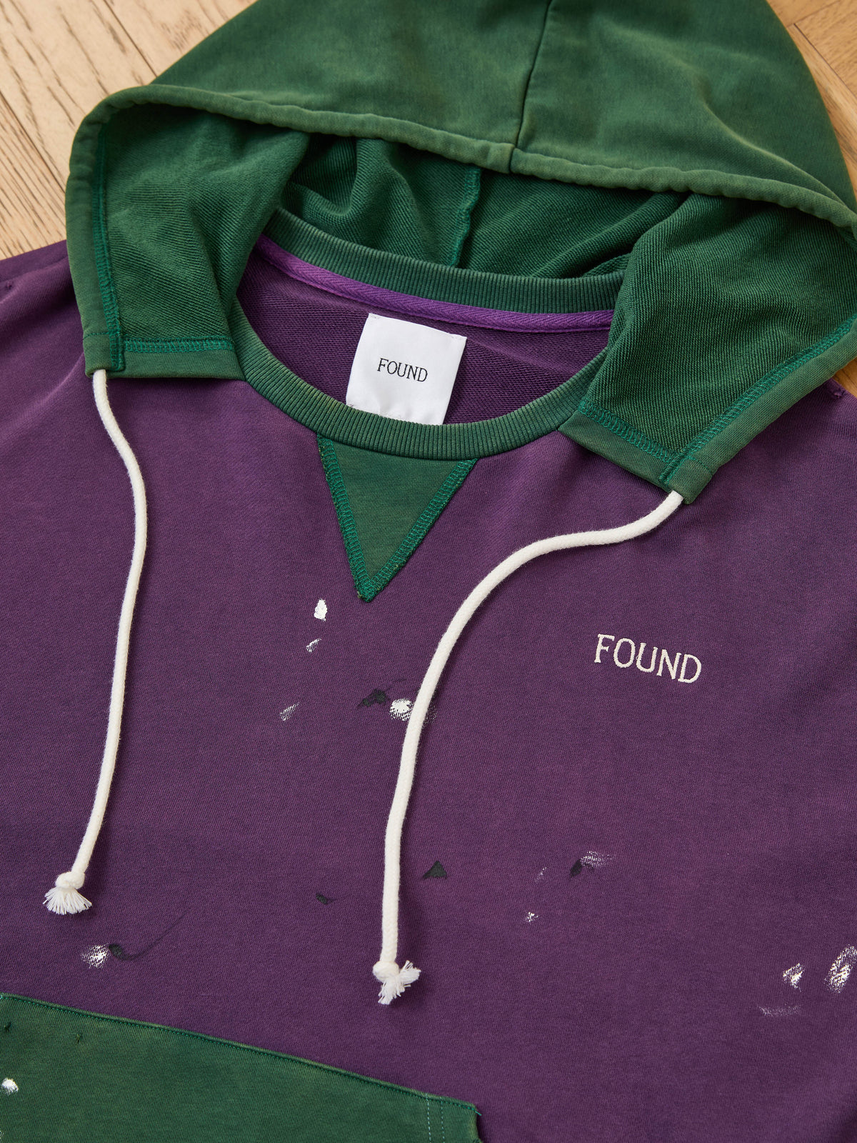 The FOUND Two Tone Painter Hoodie in purple and green with white drawstrings rests on wood, featuring small white paint stains on the front.