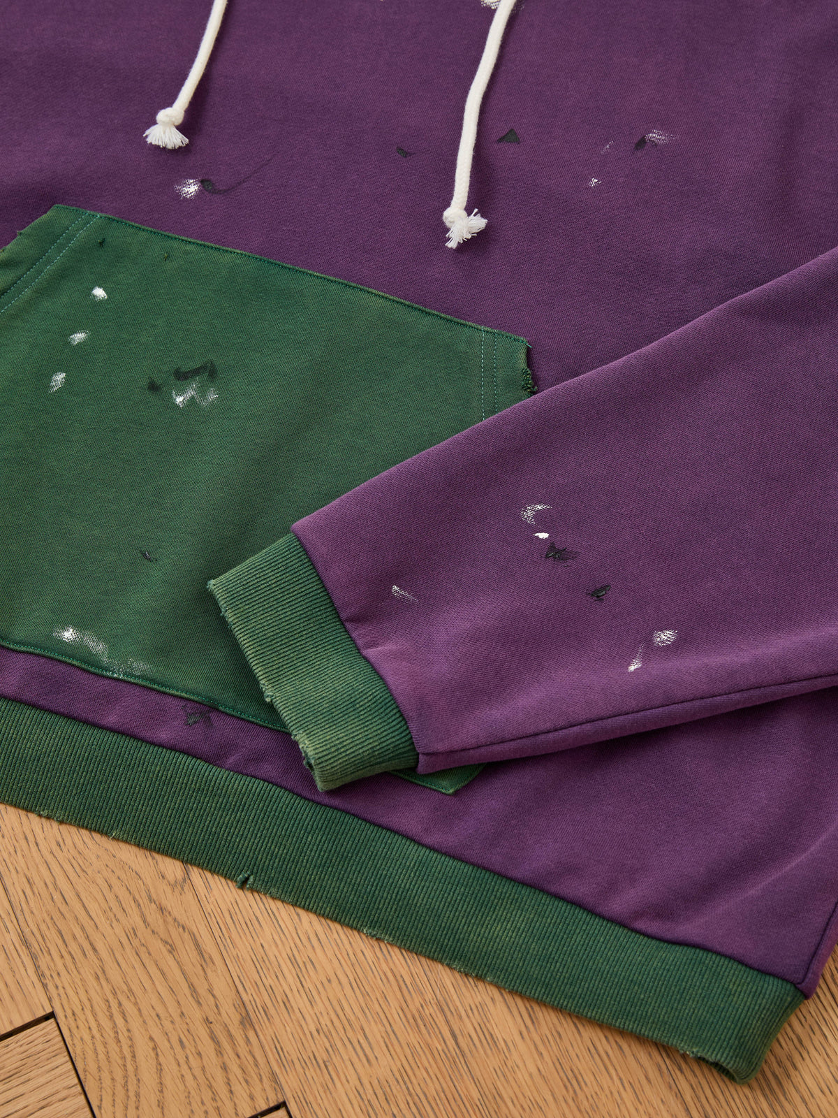 The Two Tone Painter Hoodie by FOUND, in purple and green with white drawstrings, paint splatters, and worn details, features a unisex relaxed fit.