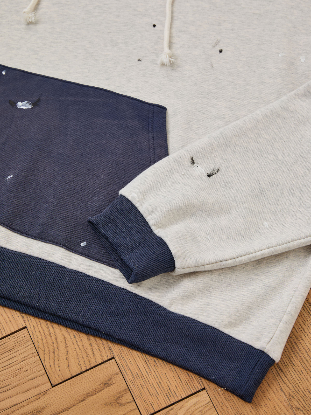 The FOUND Two-Tone Painter Hoodie features a relaxed fit in light gray with navy hem and pocket accents, highlighted by small holes and paint splatter details for a vintage-inspired look, shown laid out on a wooden floor.