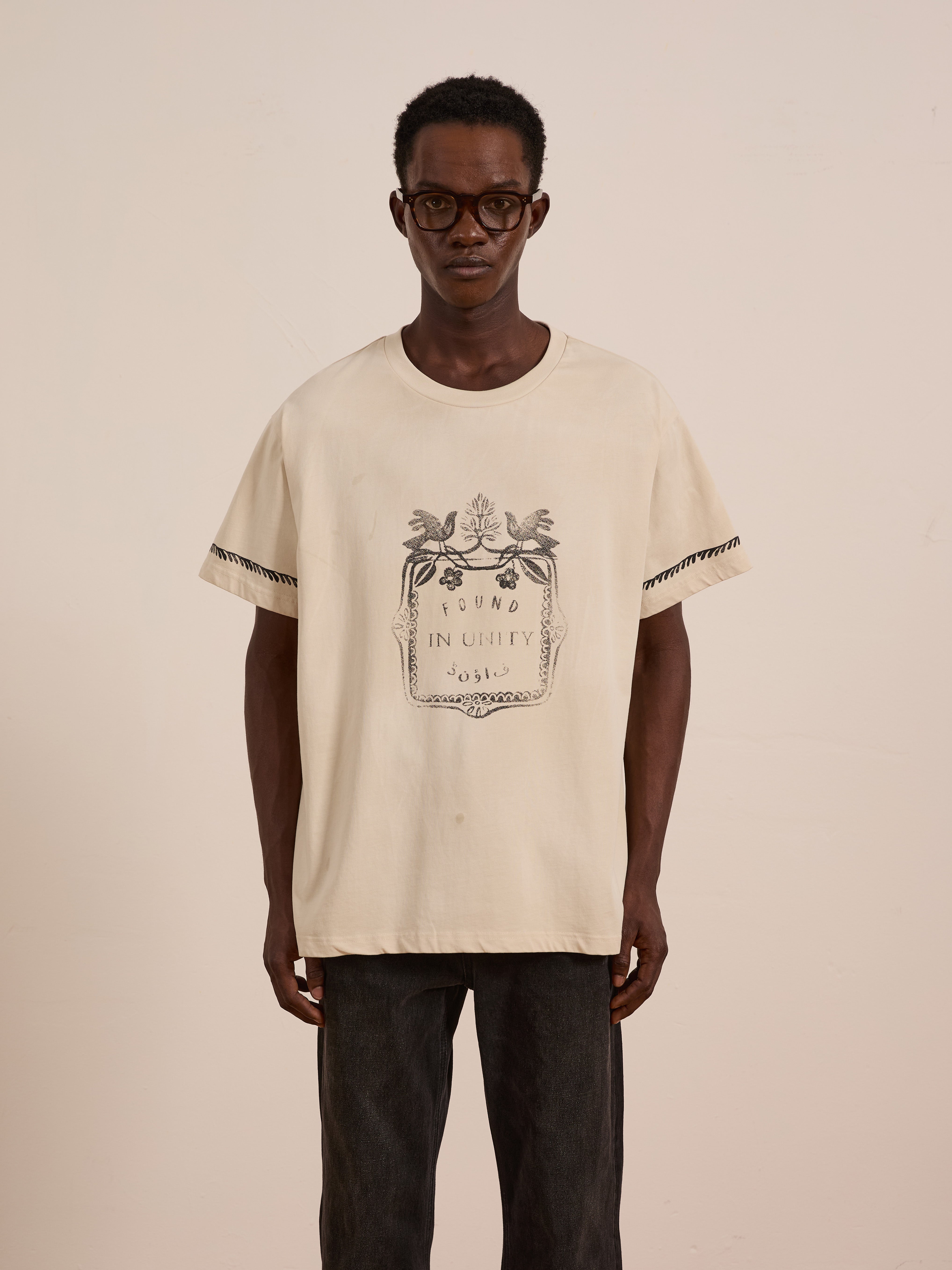 A man in glasses wears the FOUND Unity Crest Tee, a beige unisex mid-weight cotton shirt with a floral graphic and "Found In Unity" text, standing against a plain light background. This tee combines comfort and style for any occasion.
