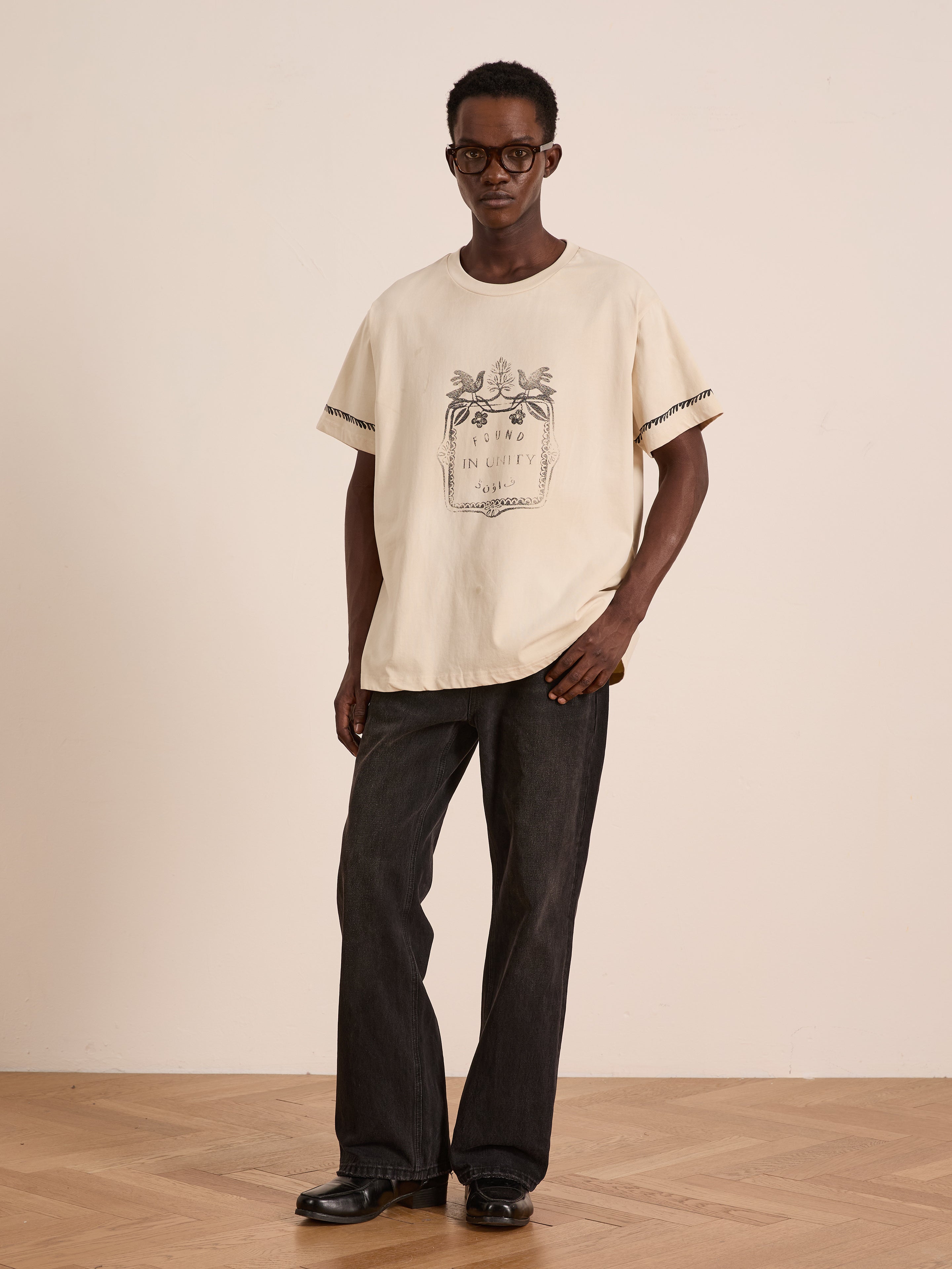 A person in glasses wears FOUND's Unity Crest Tee, a loose unisex mid-weight cotton graphic shirt, paired with dark wide-leg jeans and black shoes, standing against a plain light-colored wall.