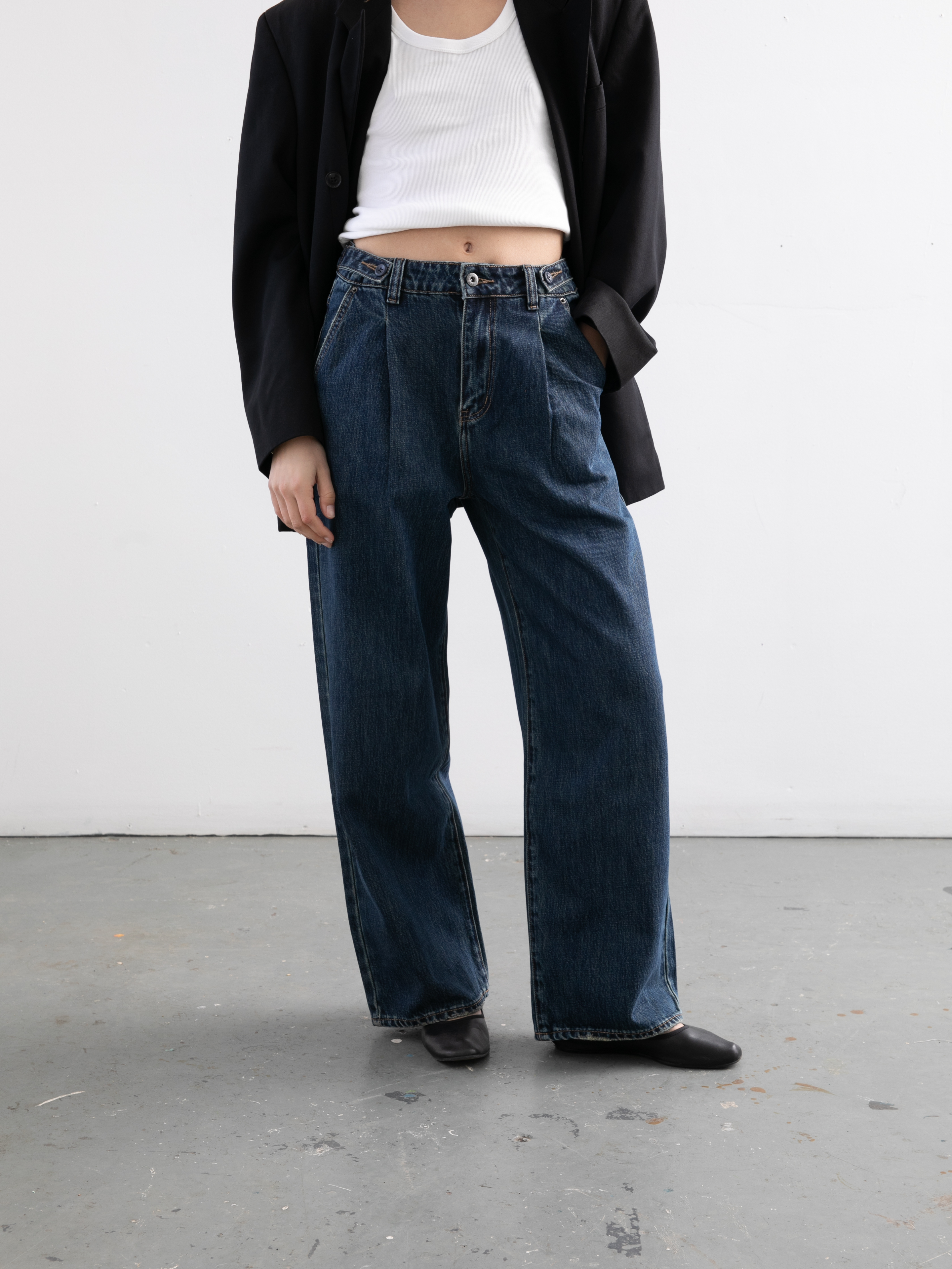 A person wears Found Pleated Relaxed Fit Jeans with a black blazer, white crop top, and black shoes, standing against a plain light wall.