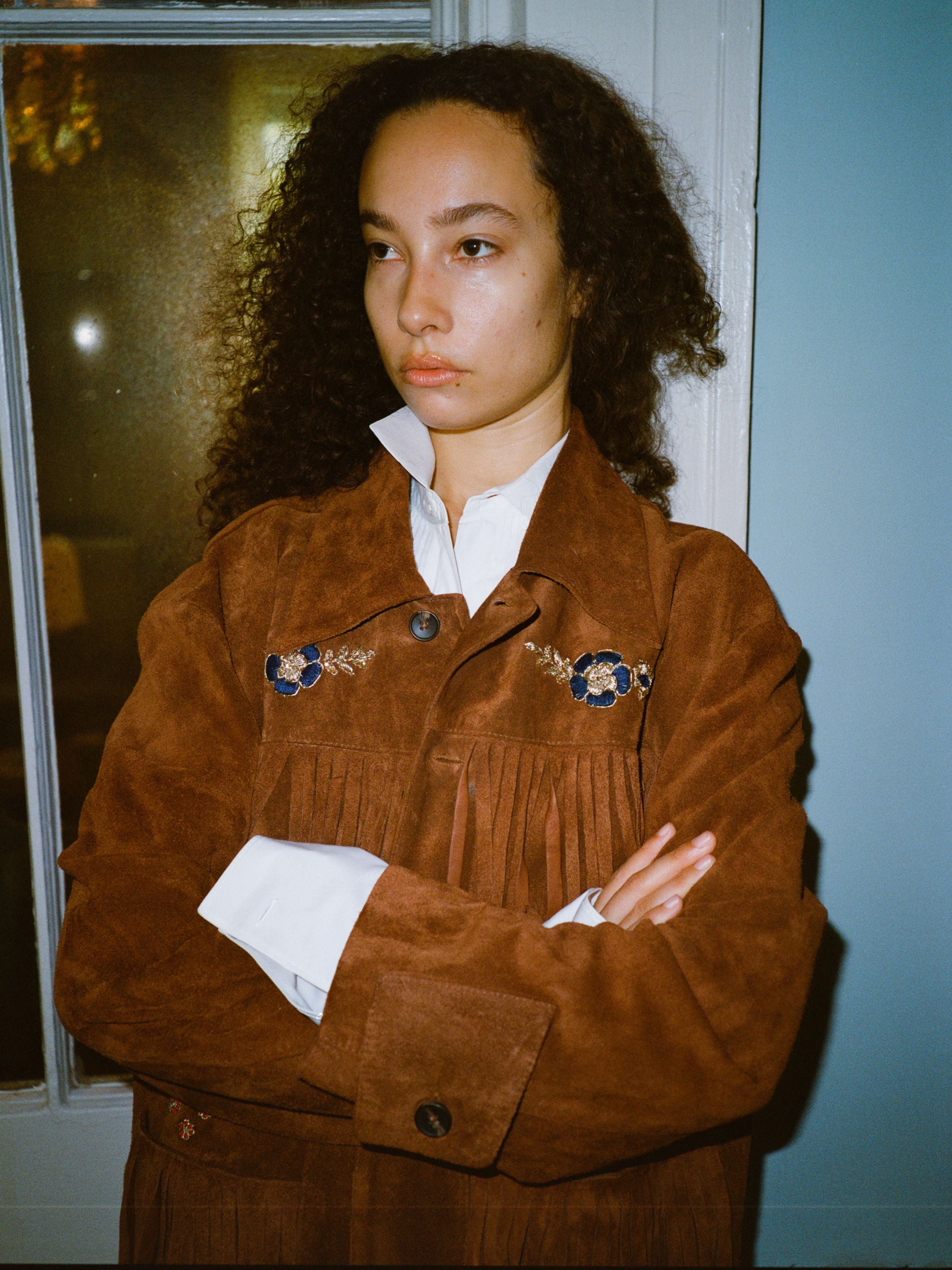 A person with curly hair stands indoors, arms crossed, wearing the FOUND Hand Embroidered Suede Fringe Jacket over a white collared shirt, facing slightly away from the camera.