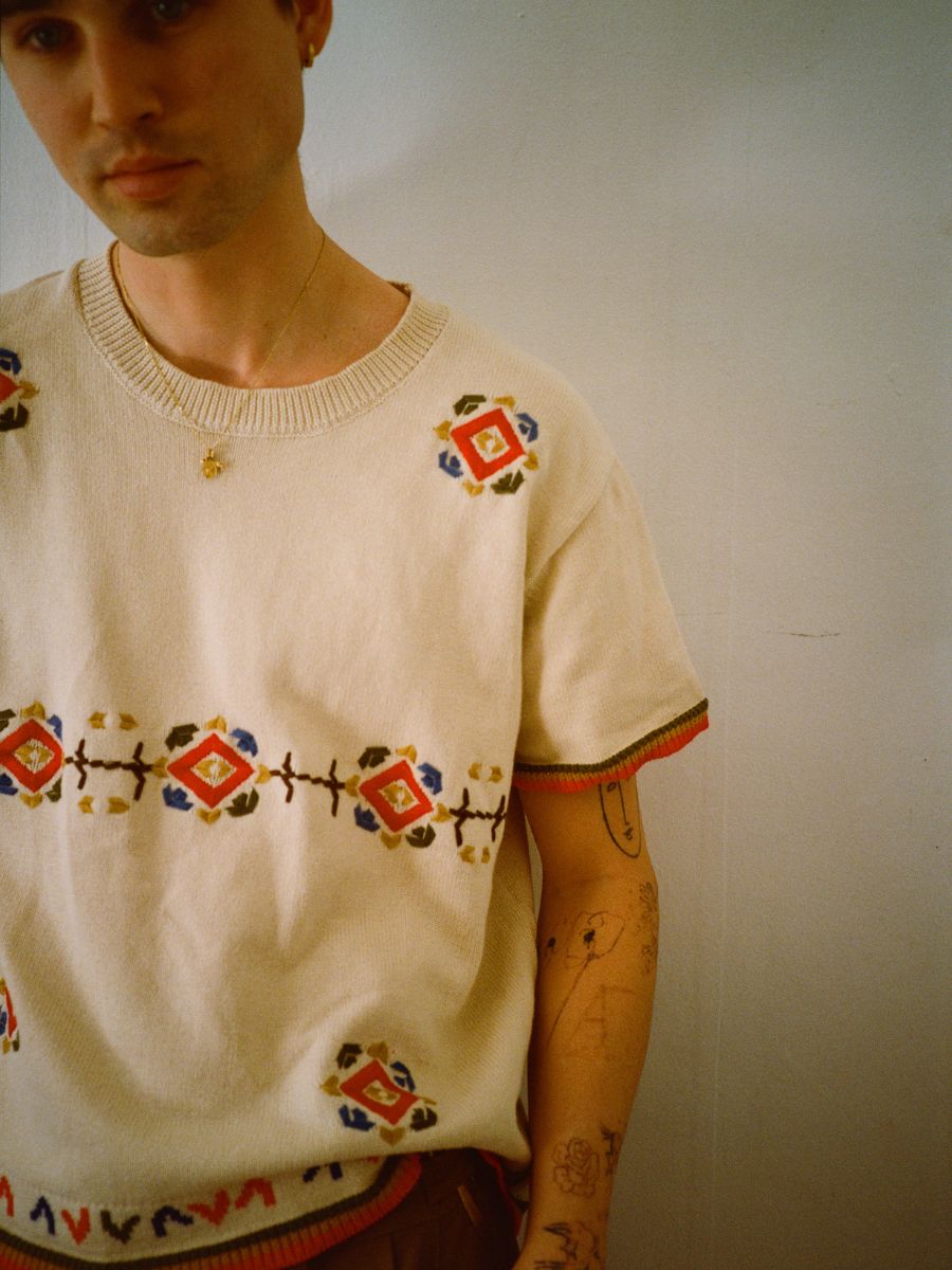 A person wears the Knit Flower Petal Tee by FOUND, featuring jacquard floral motifs, against a plain wall. The design adds vintage-inspired charm to their ensemble.
