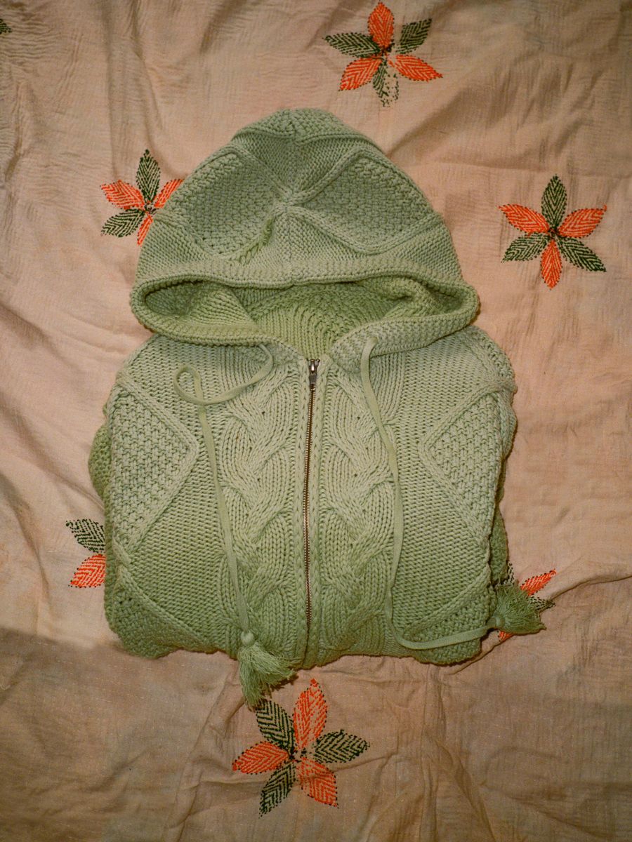 The FOUND green Zip Up Cable Knit Hoodie, made from a cotton blend yarn, is displayed on a cream fabric adorned with orange and green embroidered floral designs.