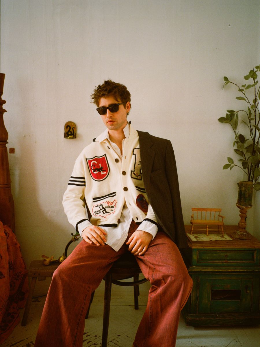 A person sits in a chair wearing FOUND sunglasses and the York Varsity Patch Collared Cardigan, paired with wide red pants and a dark coat draped over one shoulder. The vintage decor enhances the preppy aesthetic.