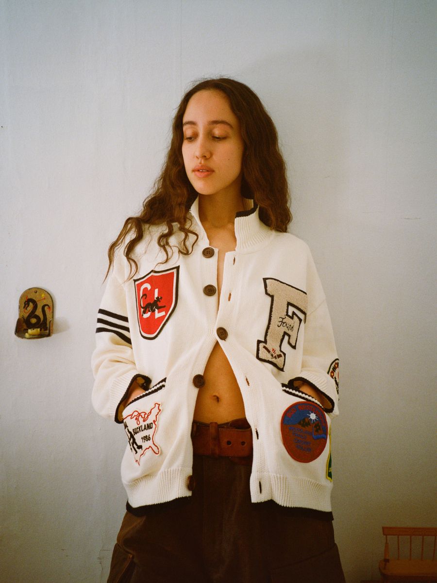 A person with long hair, fully embracing the preppy aesthetic, wears the York Varsity Patch Collared Cardigan by FOUND paired with a brown skirt. They stand against a plain wall, hands comfortably tucked in their pockets.
