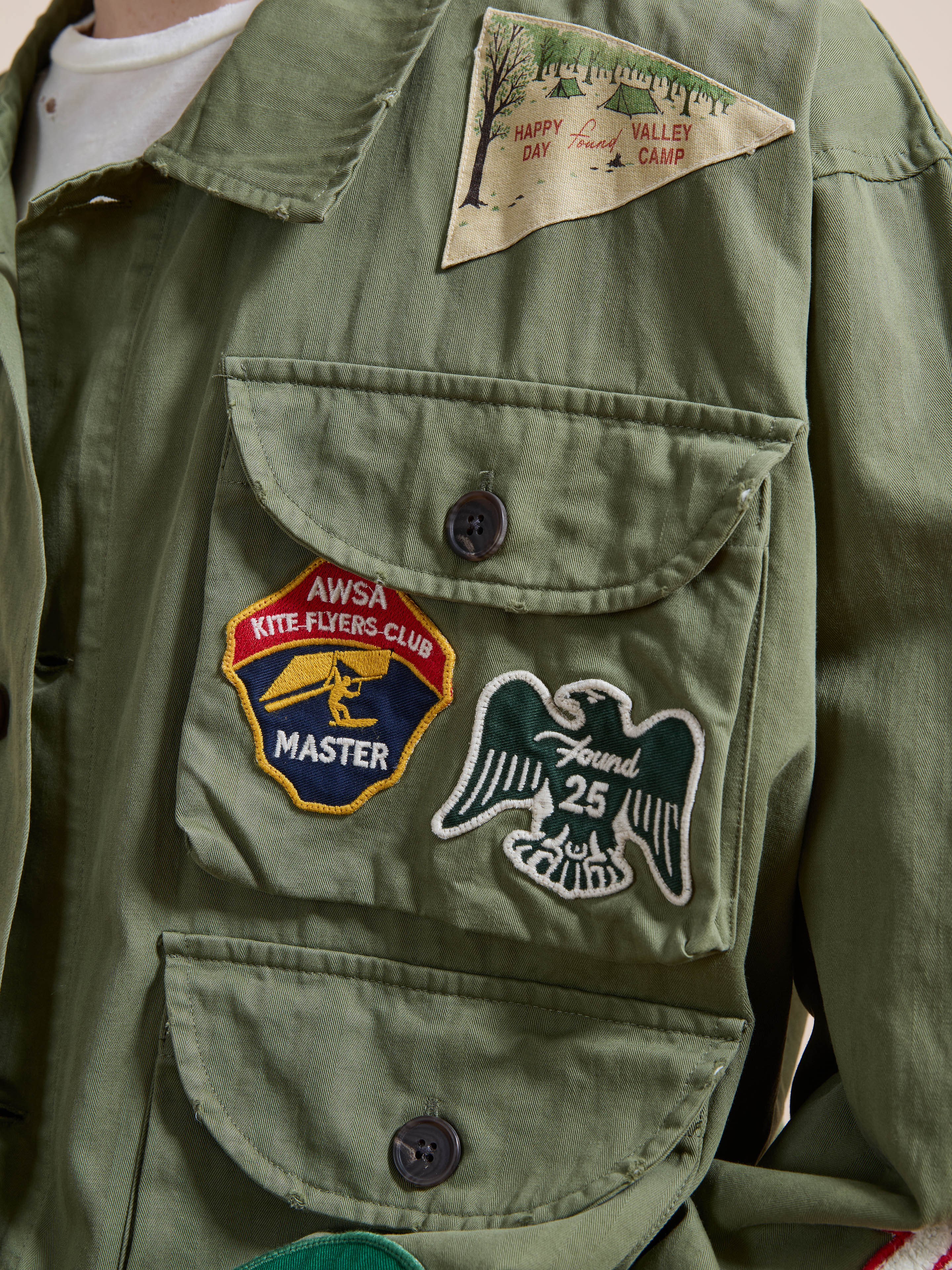 A person wears the FOUND Herringbone Utility Patchwork Jacket, featuring three embroidered patches: “AWSA Kite Flyers Club Master,” a bird with “Found 25,” and a triangle patch reading “Happy Valley Day Camp.”.