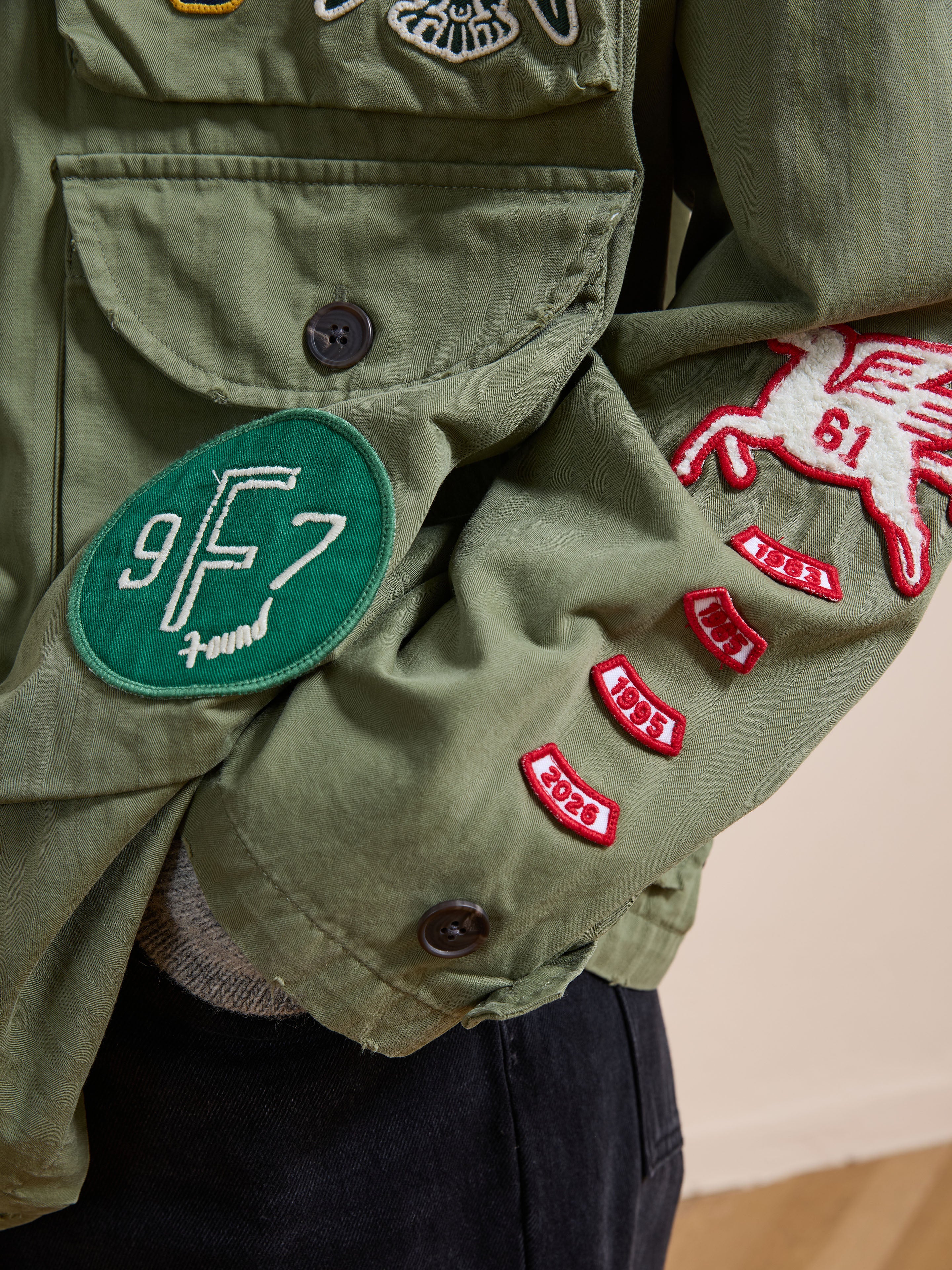 Close-up of the FOUND Herringbone Utility Patchwork Jacket in green cotton, featuring multiple embroidered patches, including a round green "9F7 Found" patch and several red date and winged horse patches.