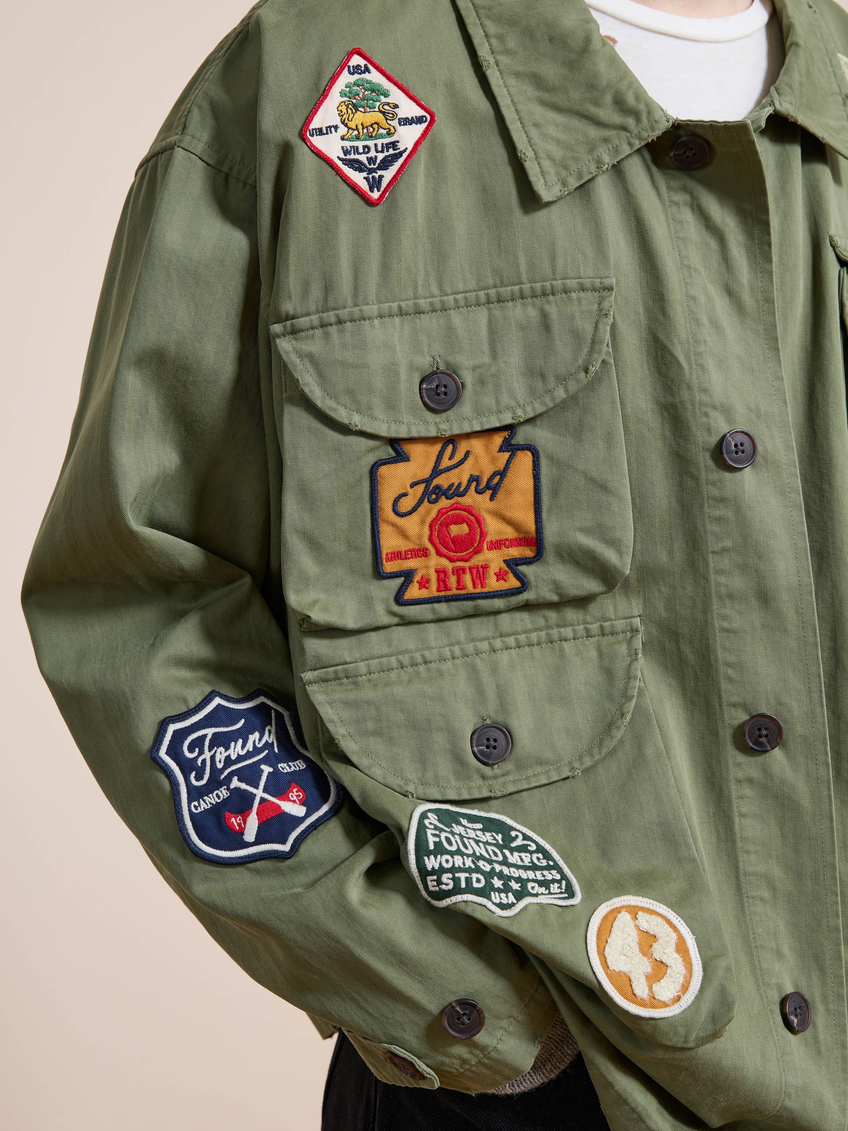 A person wearing the FOUND Herringbone Utility Patchwork Jacket, a military-inspired olive green piece decorated with colorful embroidered chest and sleeve patches.