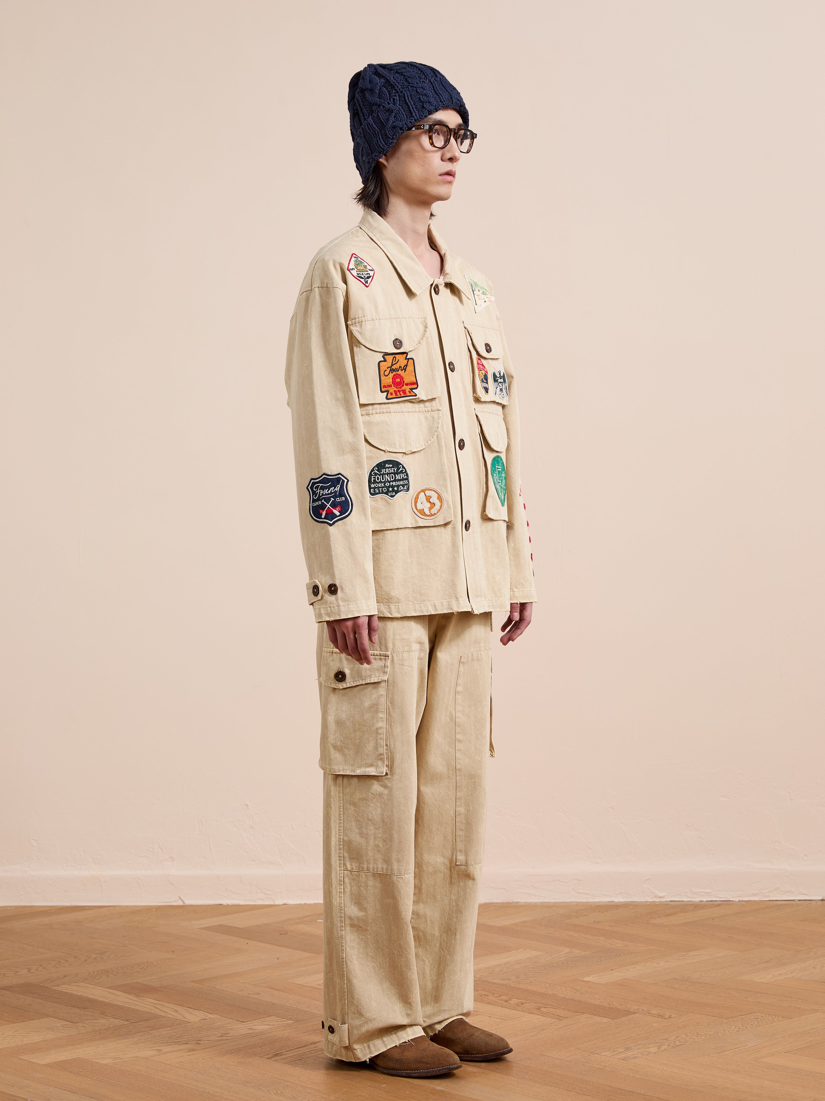 Indoors, a person in FOUND's Herringbone Utility Patchwork Jacket and matching cargo pants, plus a navy knit beanie, glasses, and brown shoes, stands on a wooden floor facing left.