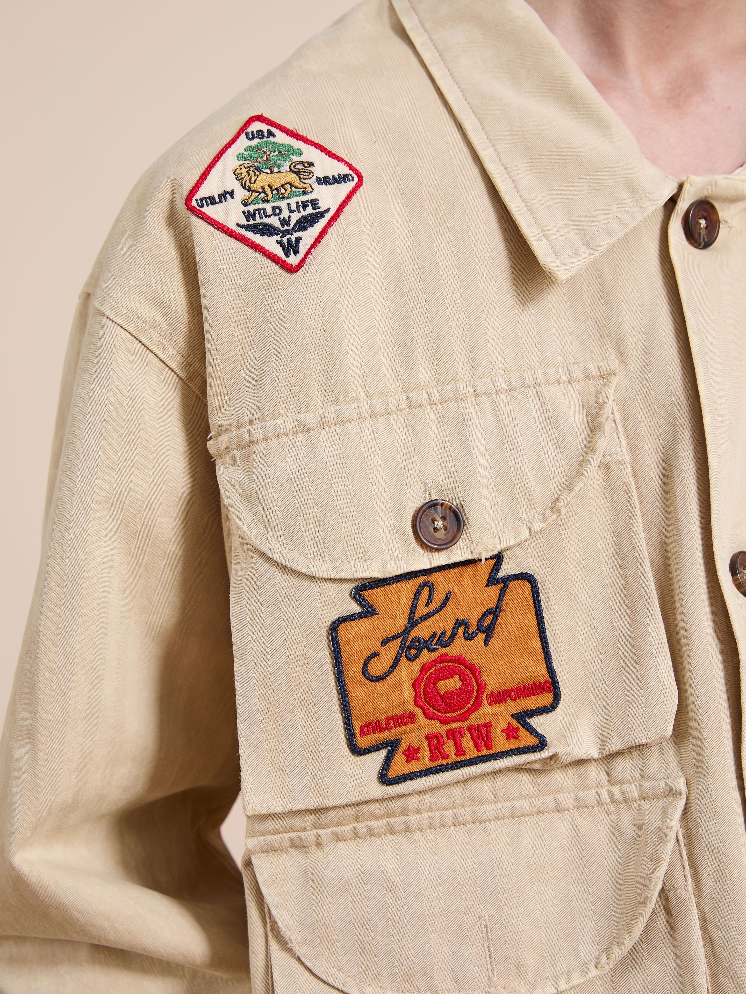 A close-up of the FOUND Herringbone Utility Patchwork Jacket shows two embroidered patches—one on the chest reading "Found Athletics RTW," and another diamond-shaped patch on the shoulder with "Wild Life" text.