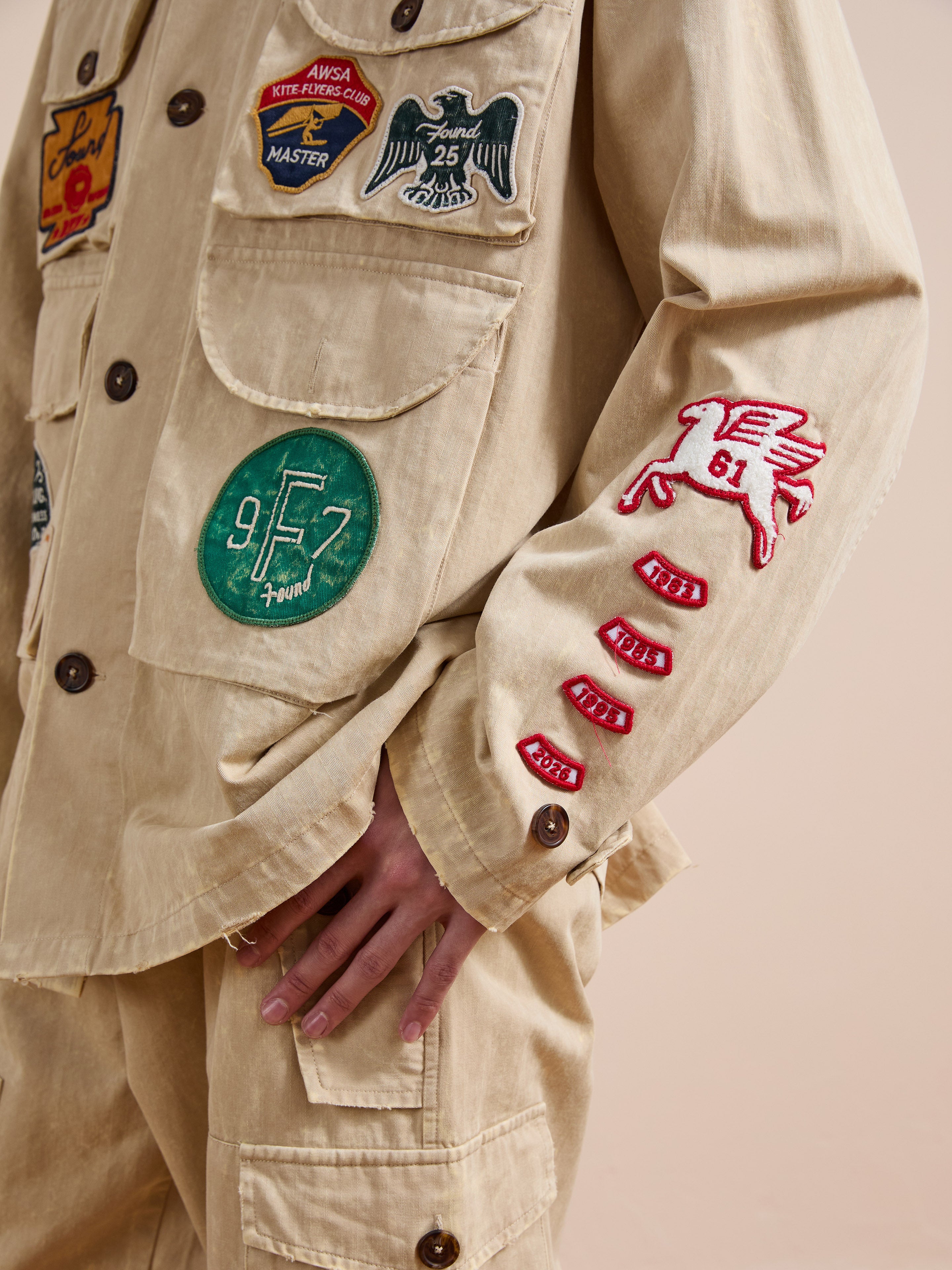 A person wearing the FOUND Herringbone Utility Patchwork Jacket, a beige vintage-style piece with multiple colorful embroidered patches, including a flying horse on the sleeve.