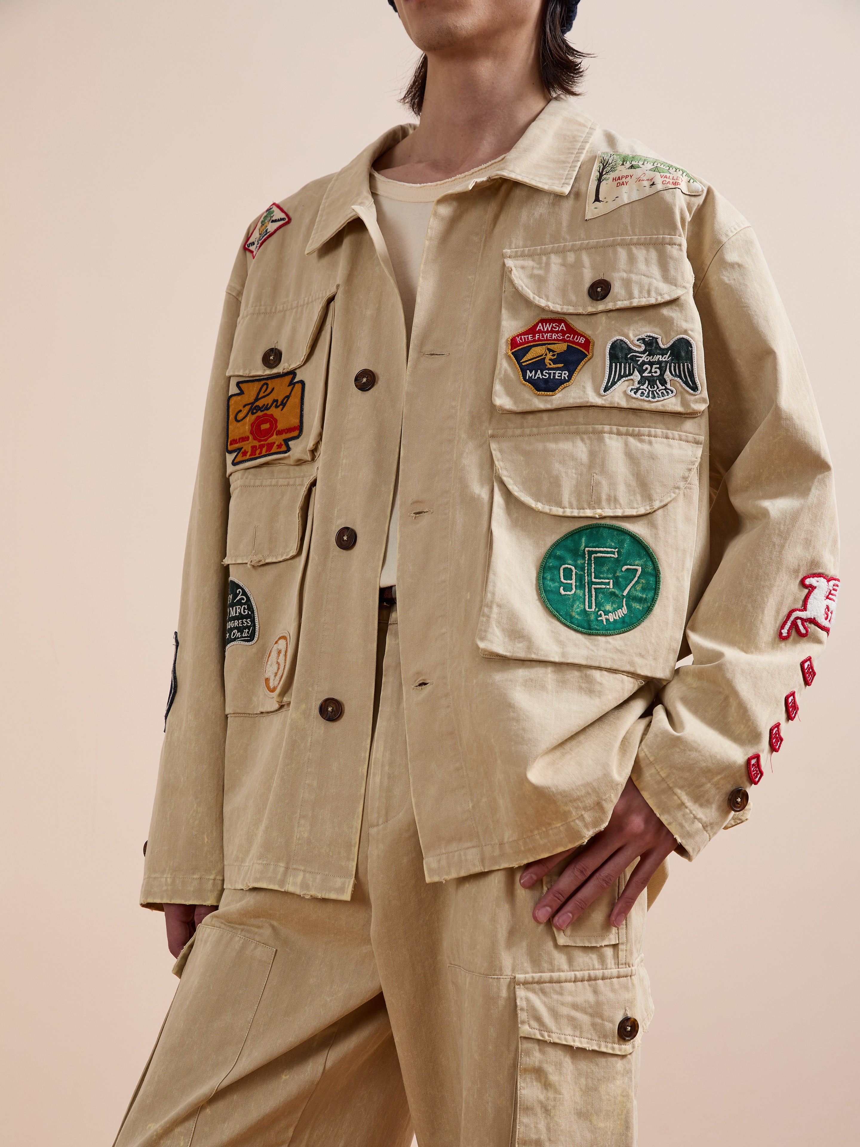 A person sports the FOUND Herringbone Utility Patchwork Jacket, featuring colorful embroidered patches on the chest, sleeves, and pockets, paired with matching pants against a plain background.