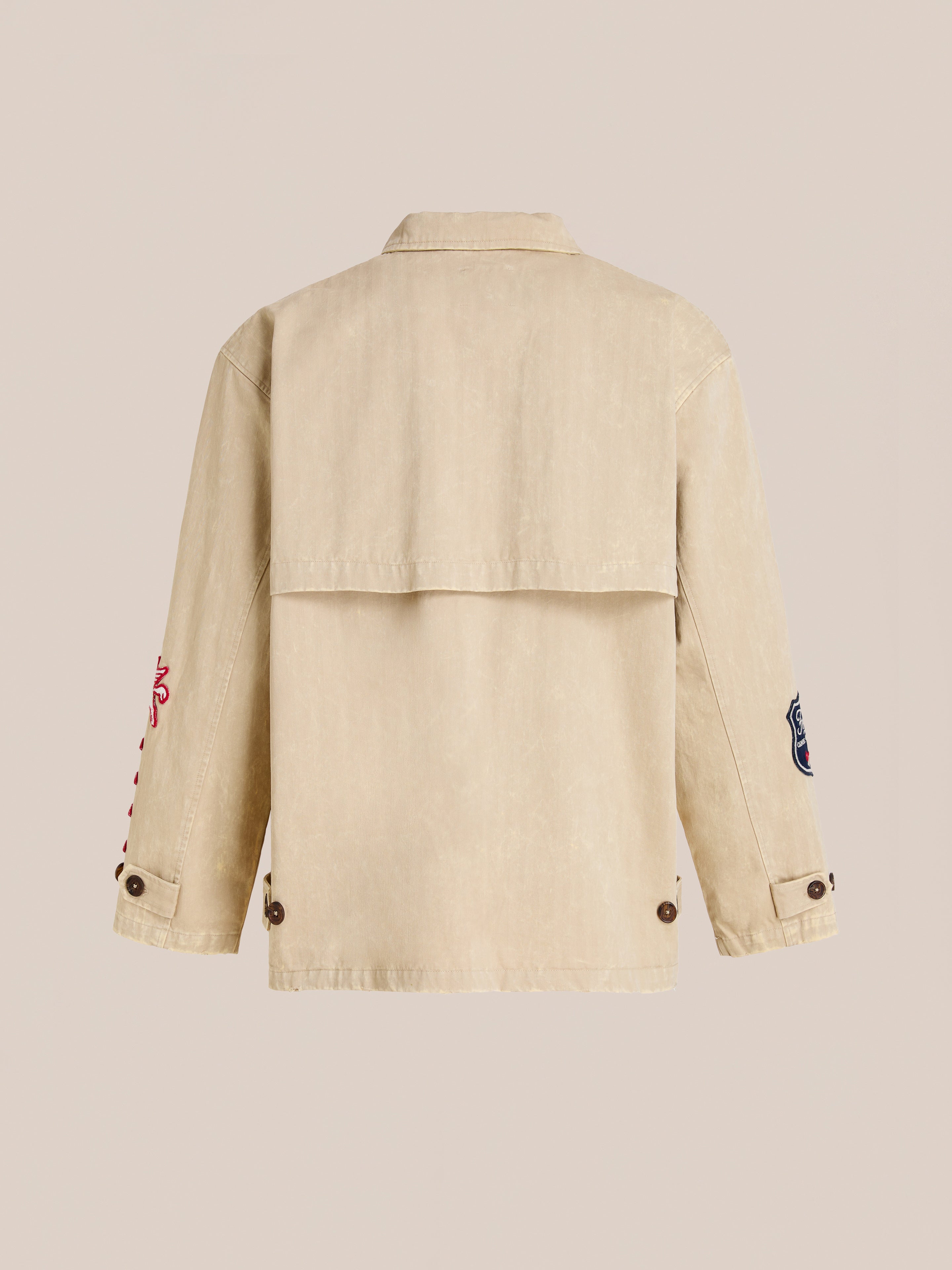 The FOUND Herringbone Utility Patchwork Jacket in beige is shown from the back, featuring long sleeves with buttoned cuffs and distinctive embroidered patches on each arm.