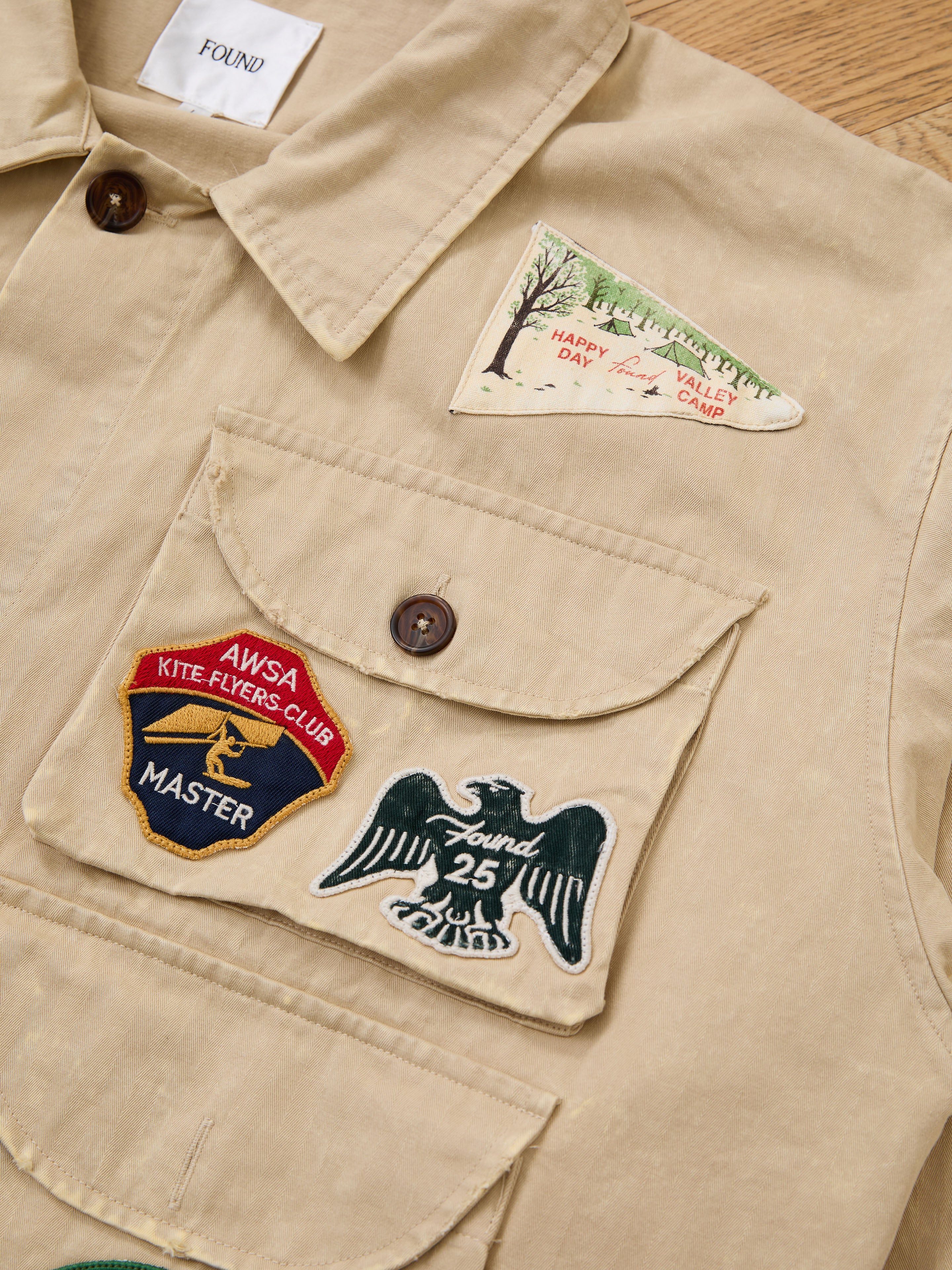 The FOUND Herringbone Utility Patchwork Jacket is a beige vintage canvas jacket with four embroidered patches: a tree scene, a kite flyers club badge, an eagle with "25," and a FOUND label on the collar.