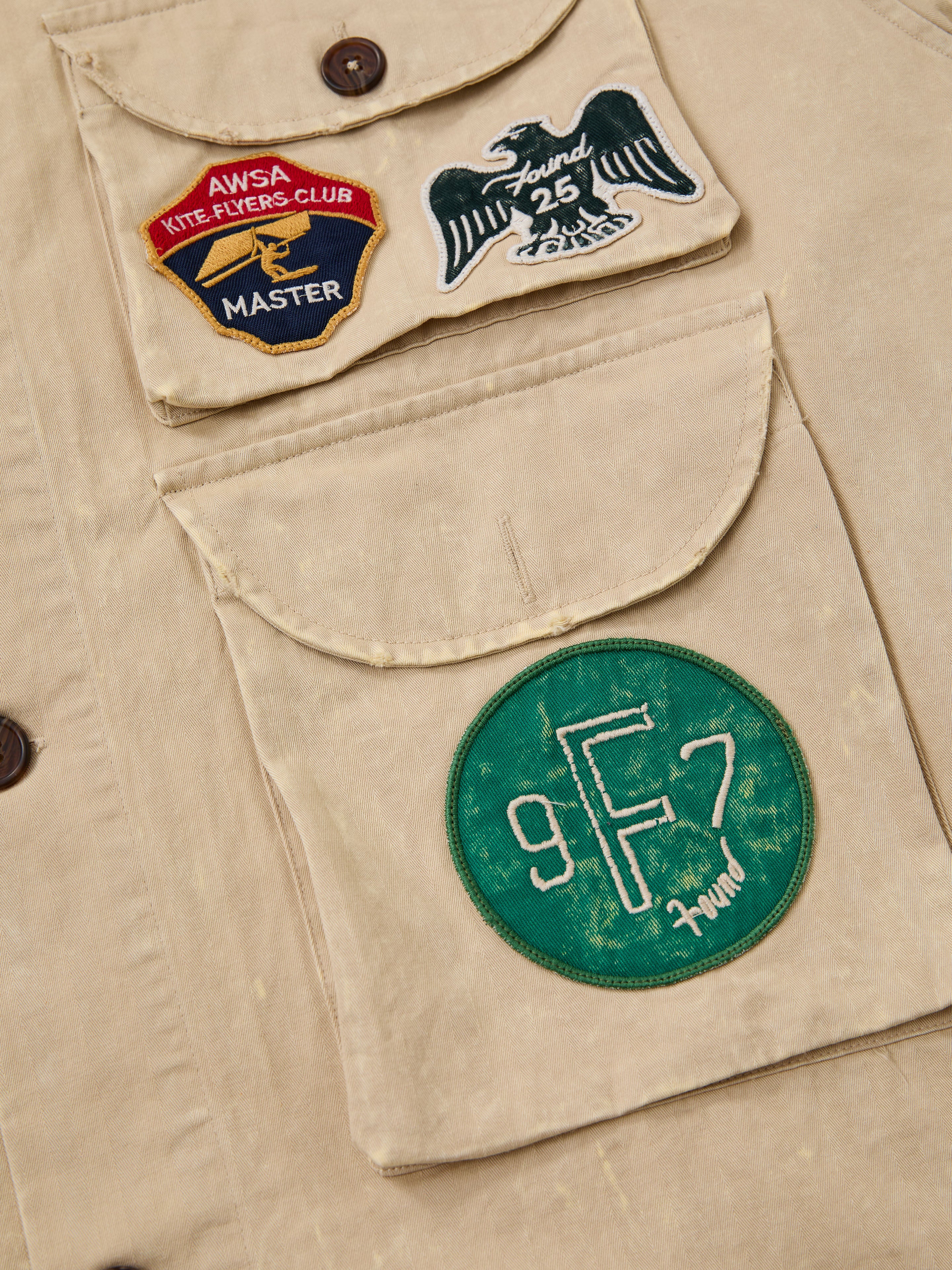 Close-up of the FOUND Herringbone Utility Patchwork Jacket, showcasing two large pockets, a green "9F7 Found" patch, eagle and AWSA Kite Flyers Club Master badges.