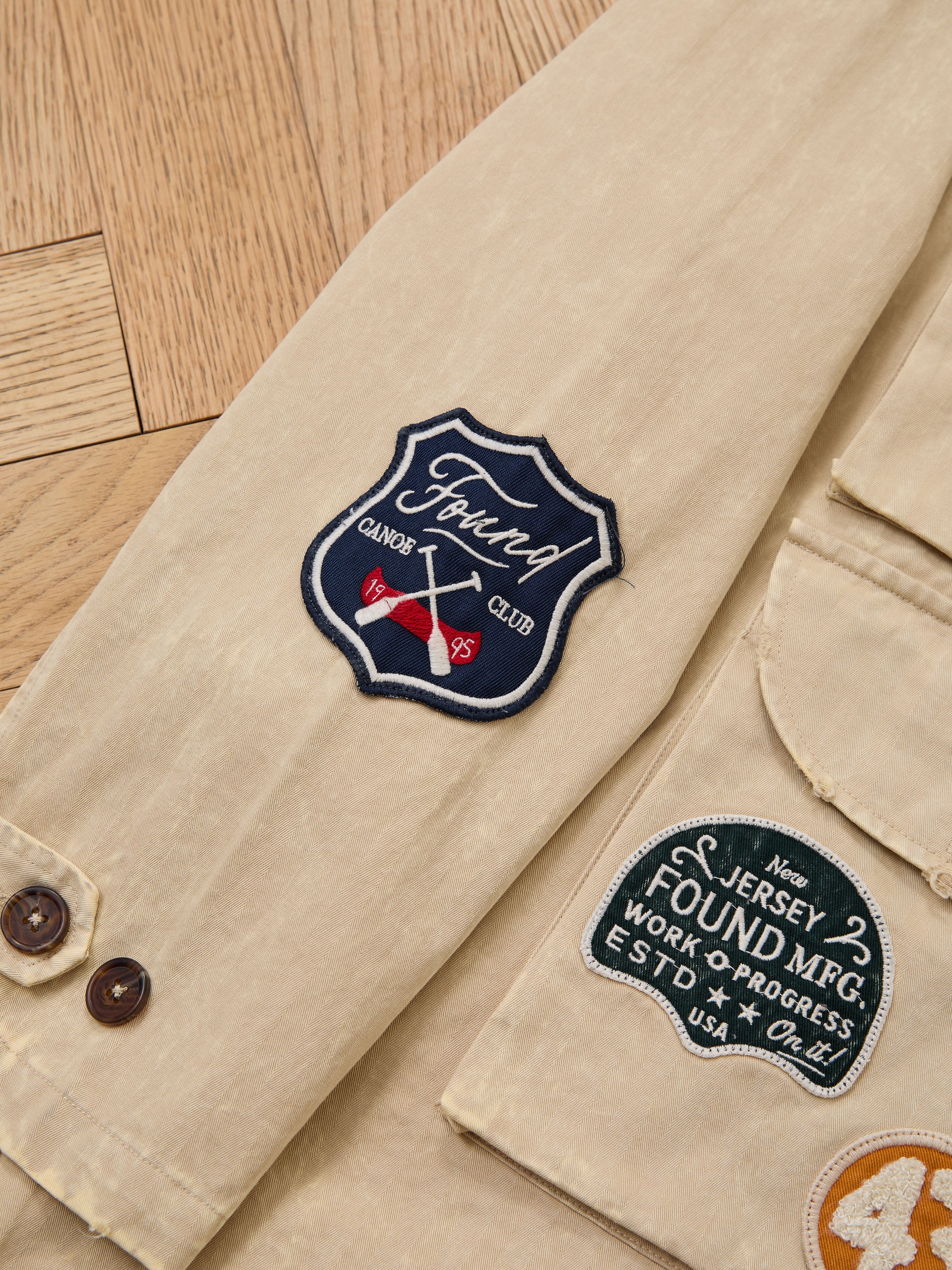The FOUND Herringbone Utility Patchwork Jacket features beige sleeves with two embroidered patches, "Found Canoe Club" and "Jersey Found MFG. Work + Progress ESTD USA." Shown against a wooden floor background.
