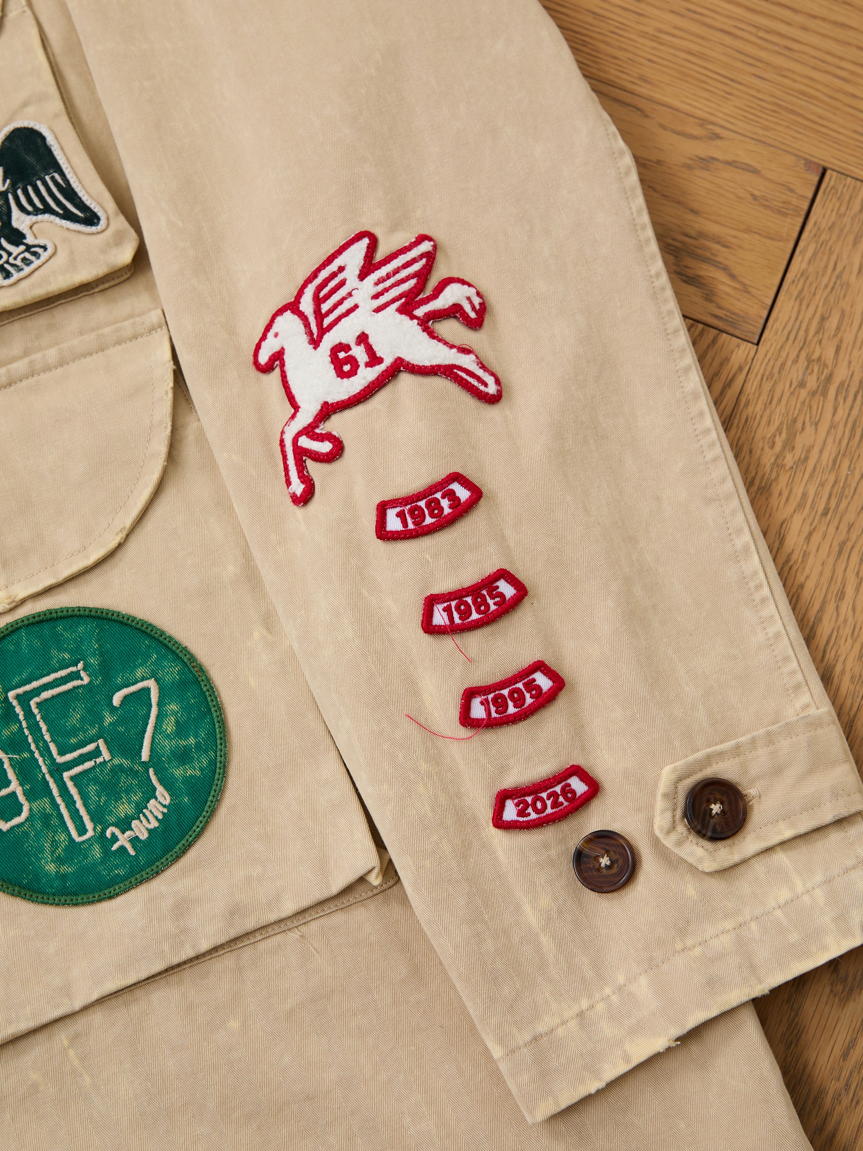 The FOUND Herringbone Utility Patchwork Jacket features a beige vintage canvas sleeve with a red "61" winged horse patch, four red year patches (1981, 1985, 1993, 2023), and additional patches on the main body. Unisex design.
