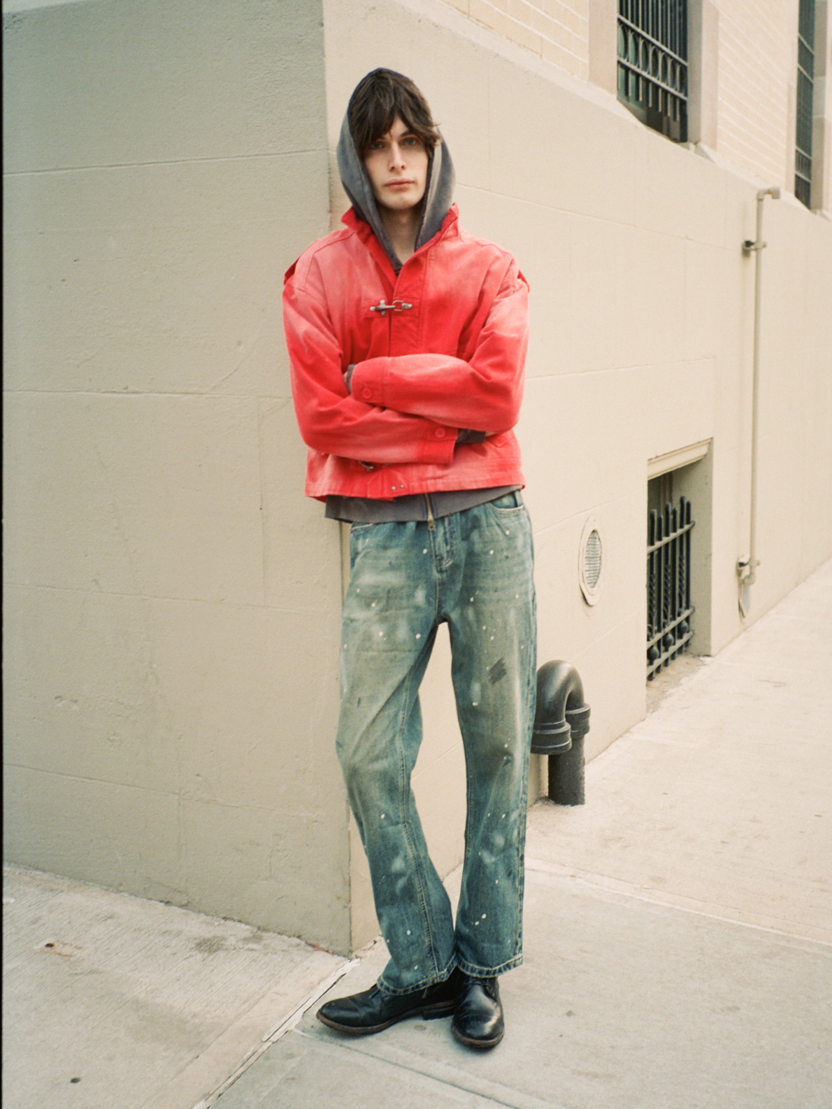 A person stands on a sidewalk with arms crossed, wearing the FOUND Utility Clasp Work Jacket over a hoodie, paint-splattered jeans, and black shoes, leaning against a light-colored building wall.