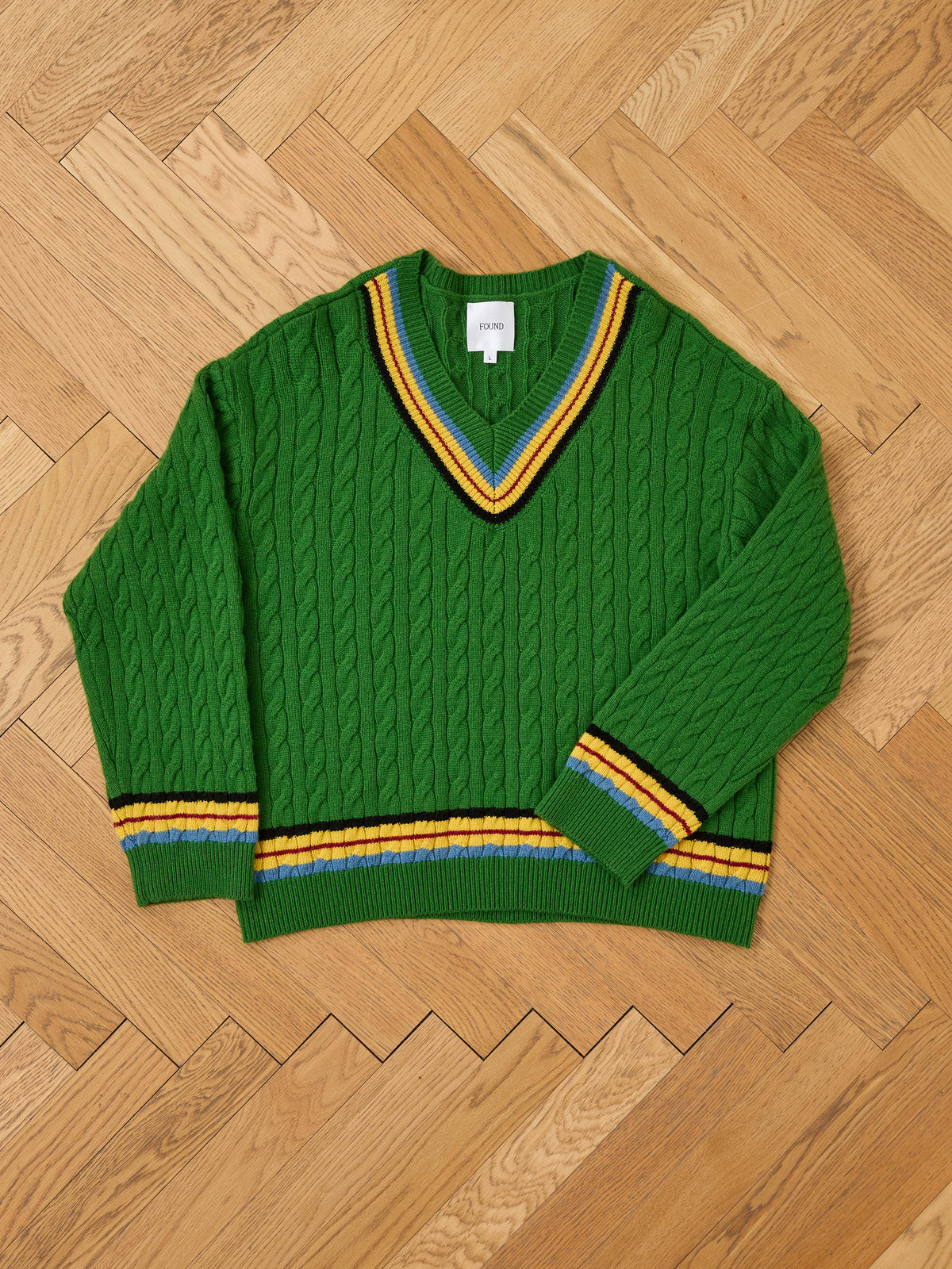 The FOUND V-Neck Cricket Sweater in green features yellow, blue, and white striped trim on the collar, cuffs, and hem, shown flat on a wooden floor.