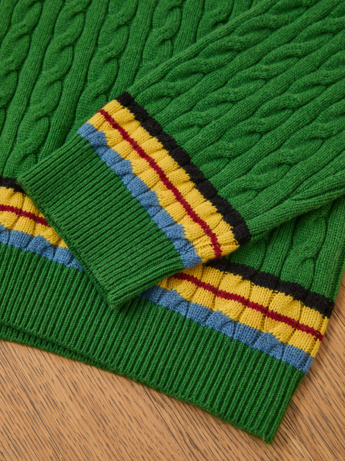 FOUND V-Neck Cricket Sweater: green cable knit with yellow, blue, red, and black striped ribbed cuffs and hem, unisex style shown on a wooden surface.