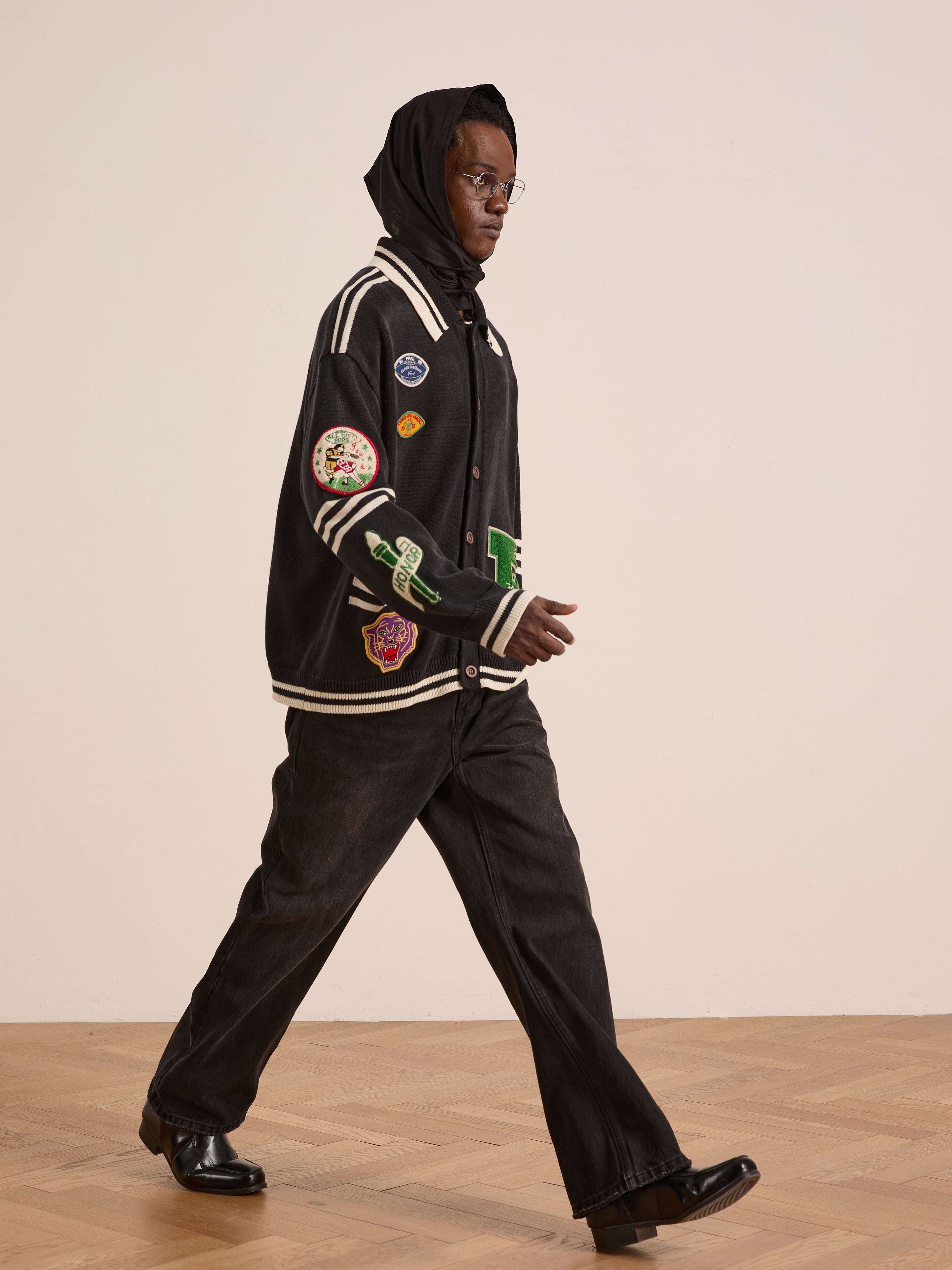 A person wearing FOUND’s Varsity Patch Collared Cardigan, black hood, glasses, black jeans, and shoes walks indoors on a wooden floor, exuding the timeless appeal of collegiate style.