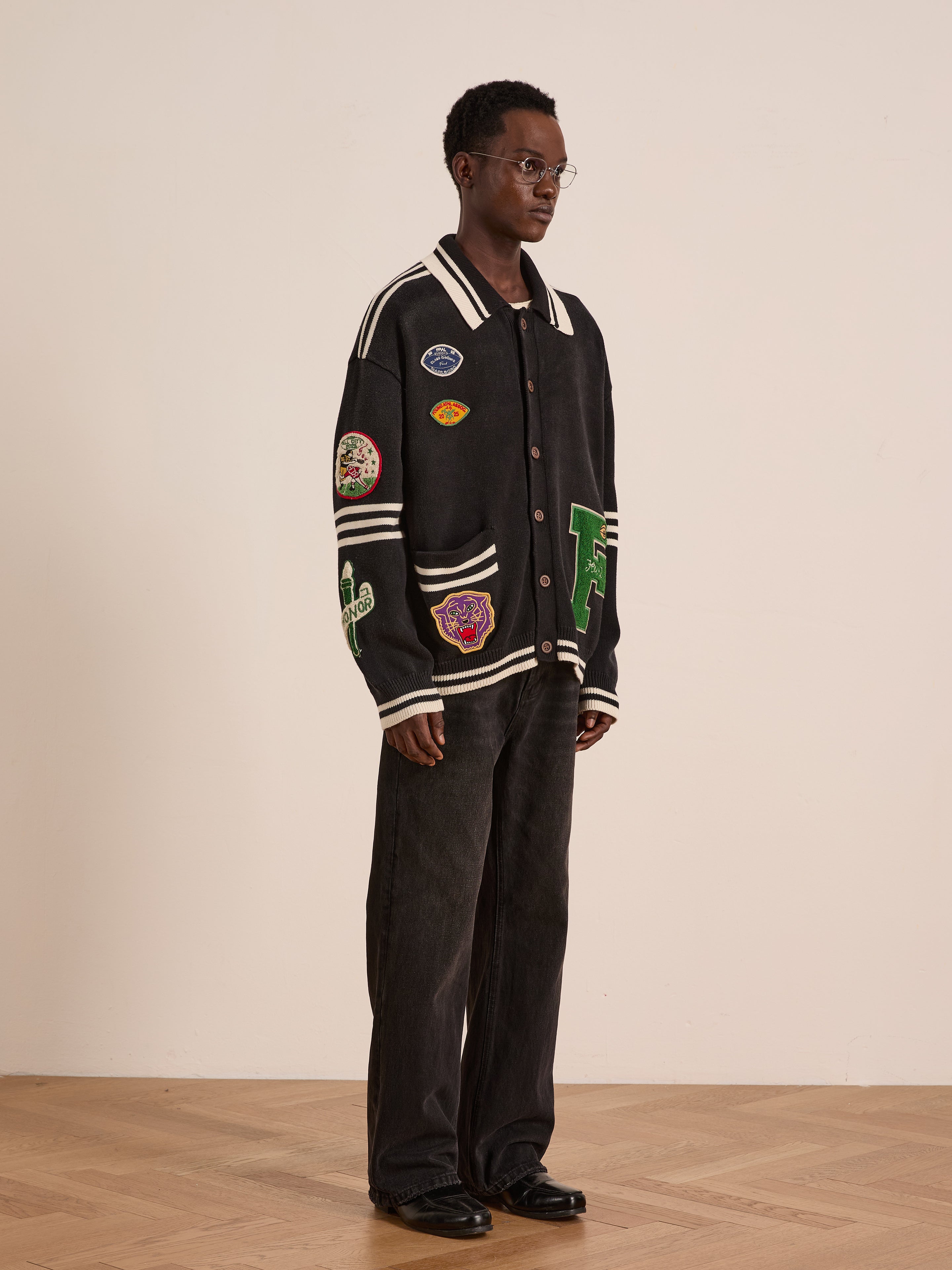 A person stands indoors against a plain background wearing the FOUND Varsity Patch Collared Cardigan with various patches, paired with black wide-leg pants and black shoes.