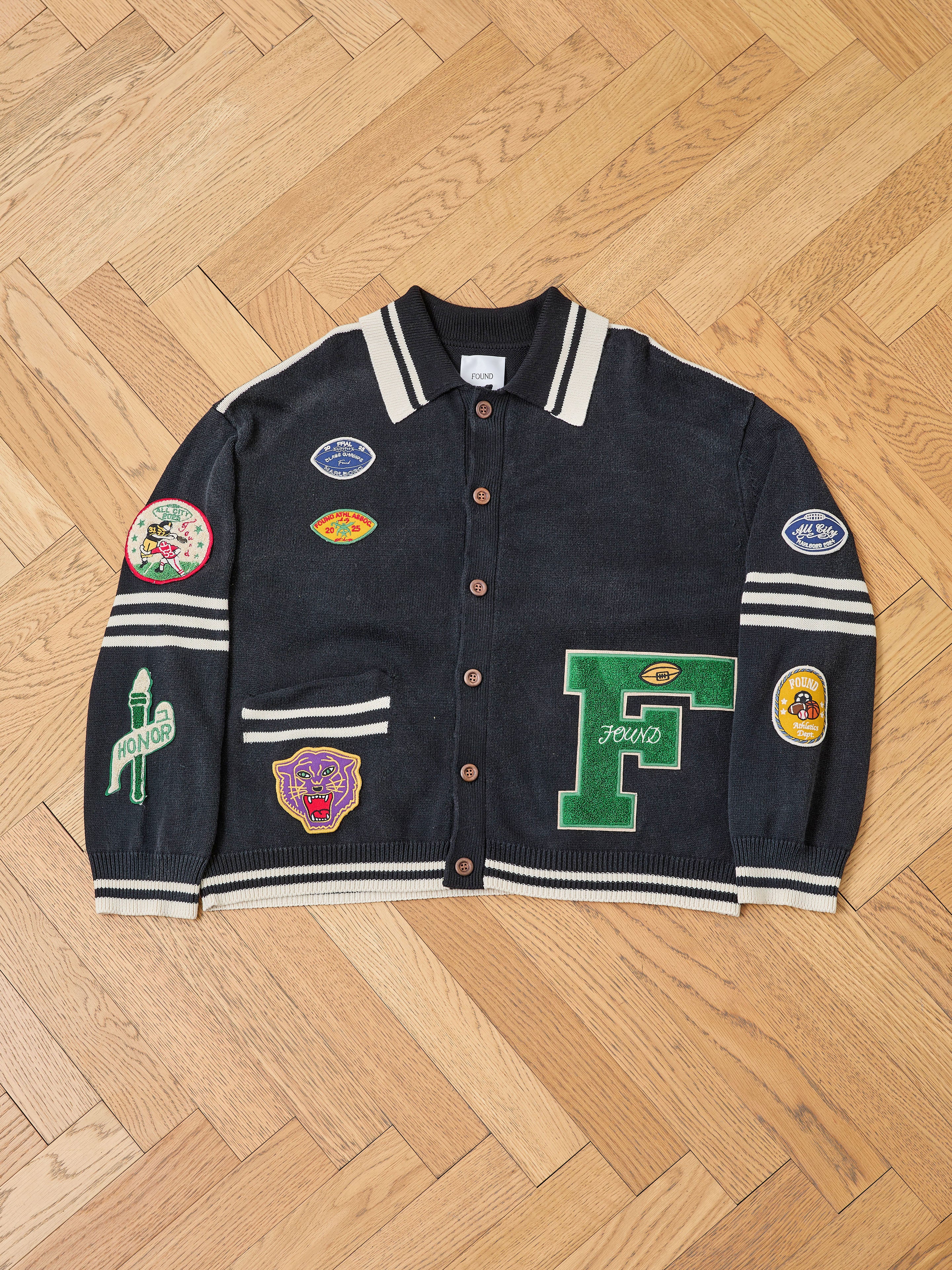 The FOUND Varsity Patch Collared Cardigan in black cotton knit, featuring white stripes and colorful chenille patches, is laid flat on a wooden herringbone floor.