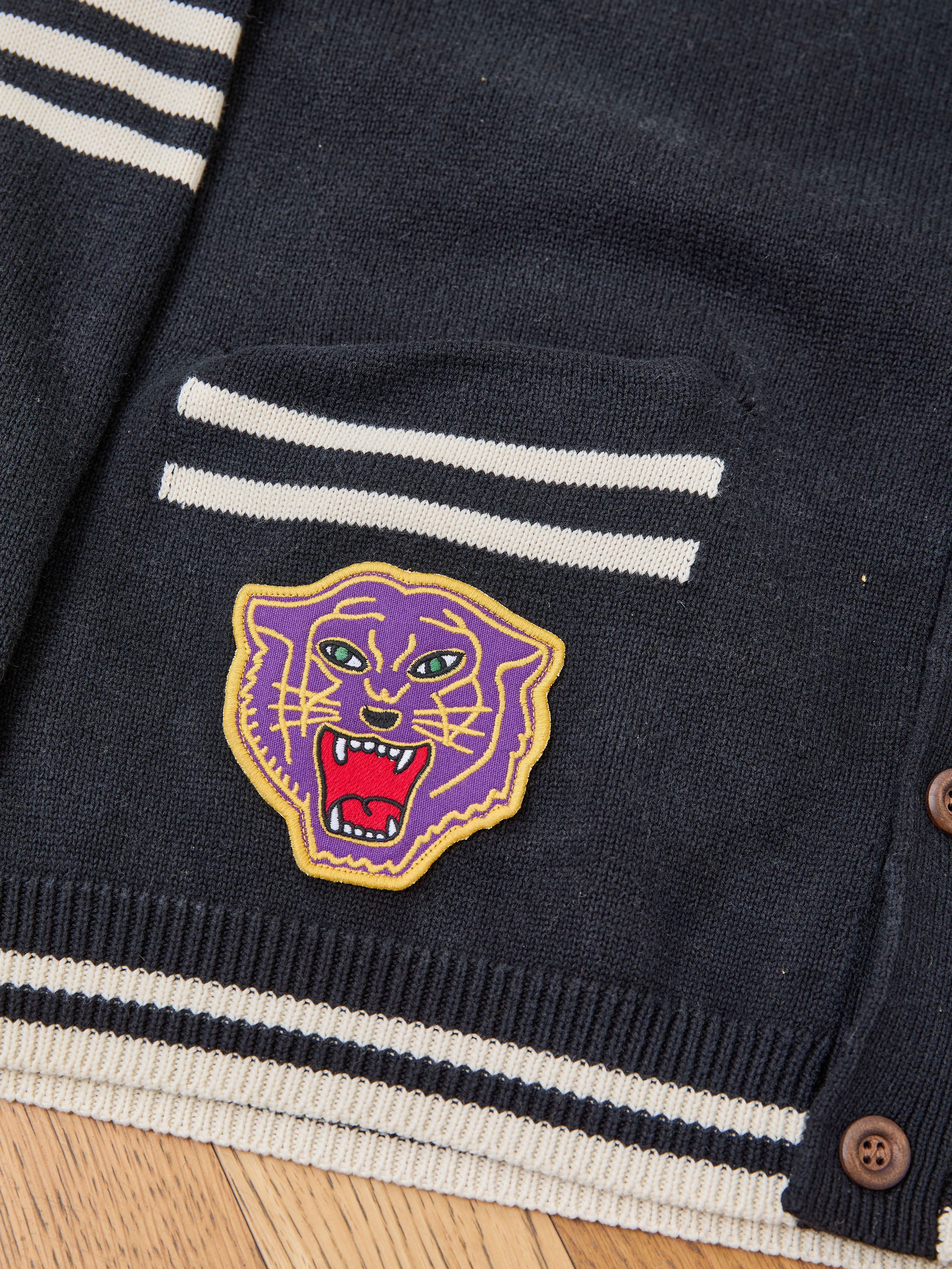 Close-up of FOUND Varsity Patch Collared Cardigan in navy blue cotton knit, featuring white stripes, brown buttons, and a chenille purple and yellow roaring panther patch above the pocket.
