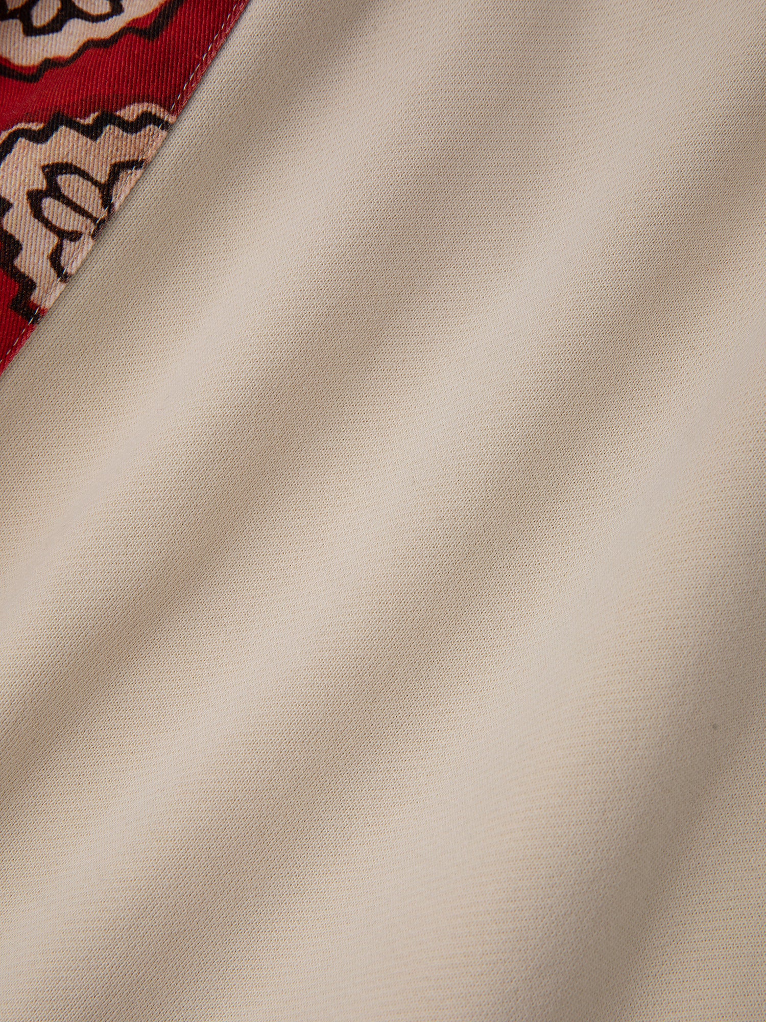 A detailed view of the Tapestry Patchwork Zip Up Hoodie by FOUND, showcasing its beige fabric intricately woven with South Asian tapestry patterns and highlighted by a red patterned border at the corner.