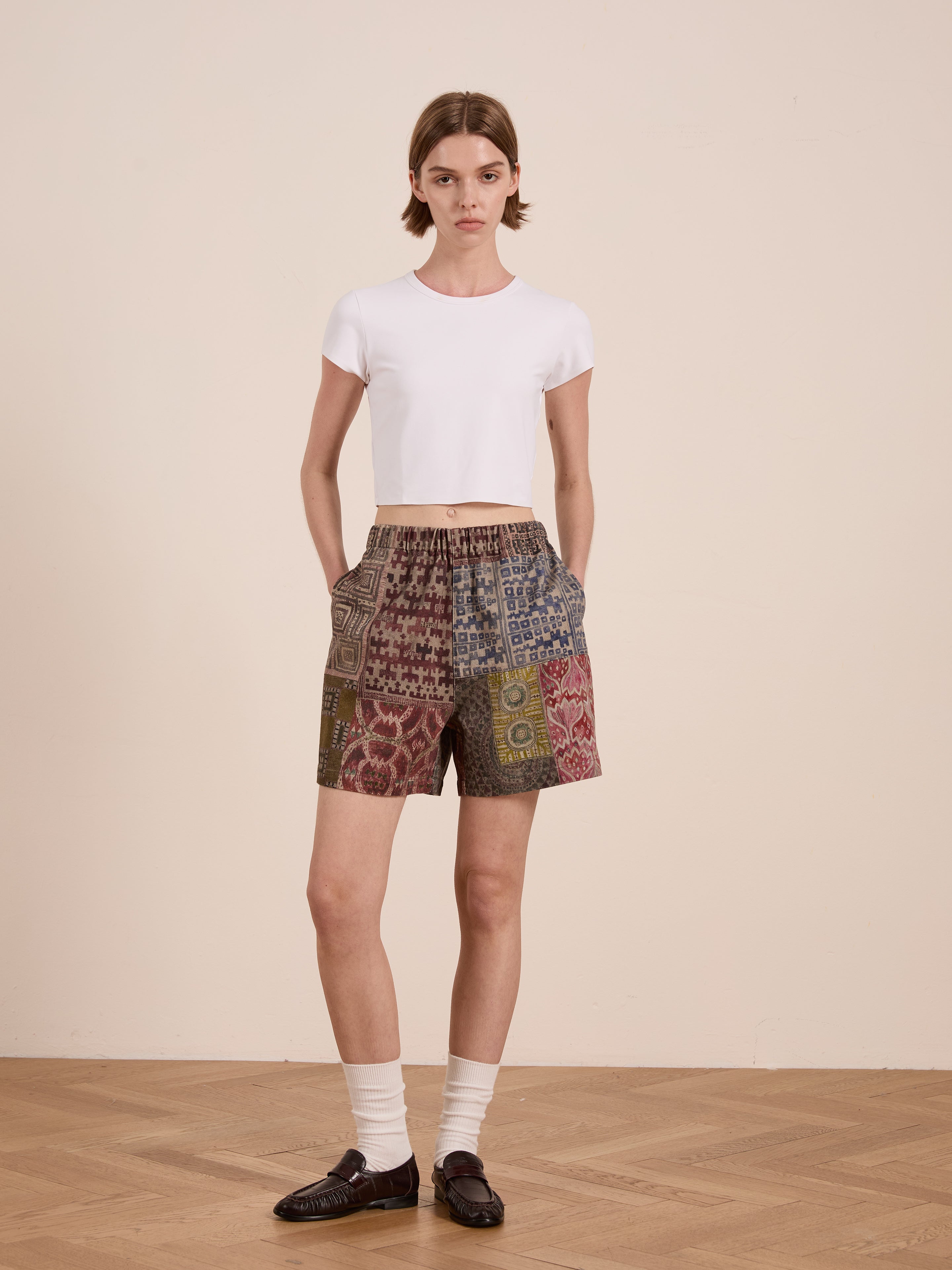 Person standing indoors wearing a FOUND white t-shirt and Watercolor Mosaic Work Shorts, highlighted by vintage-inspired patterns. Hands in pockets, socks, and loafers complete the relaxed-fit ensemble against a neutral backdrop.