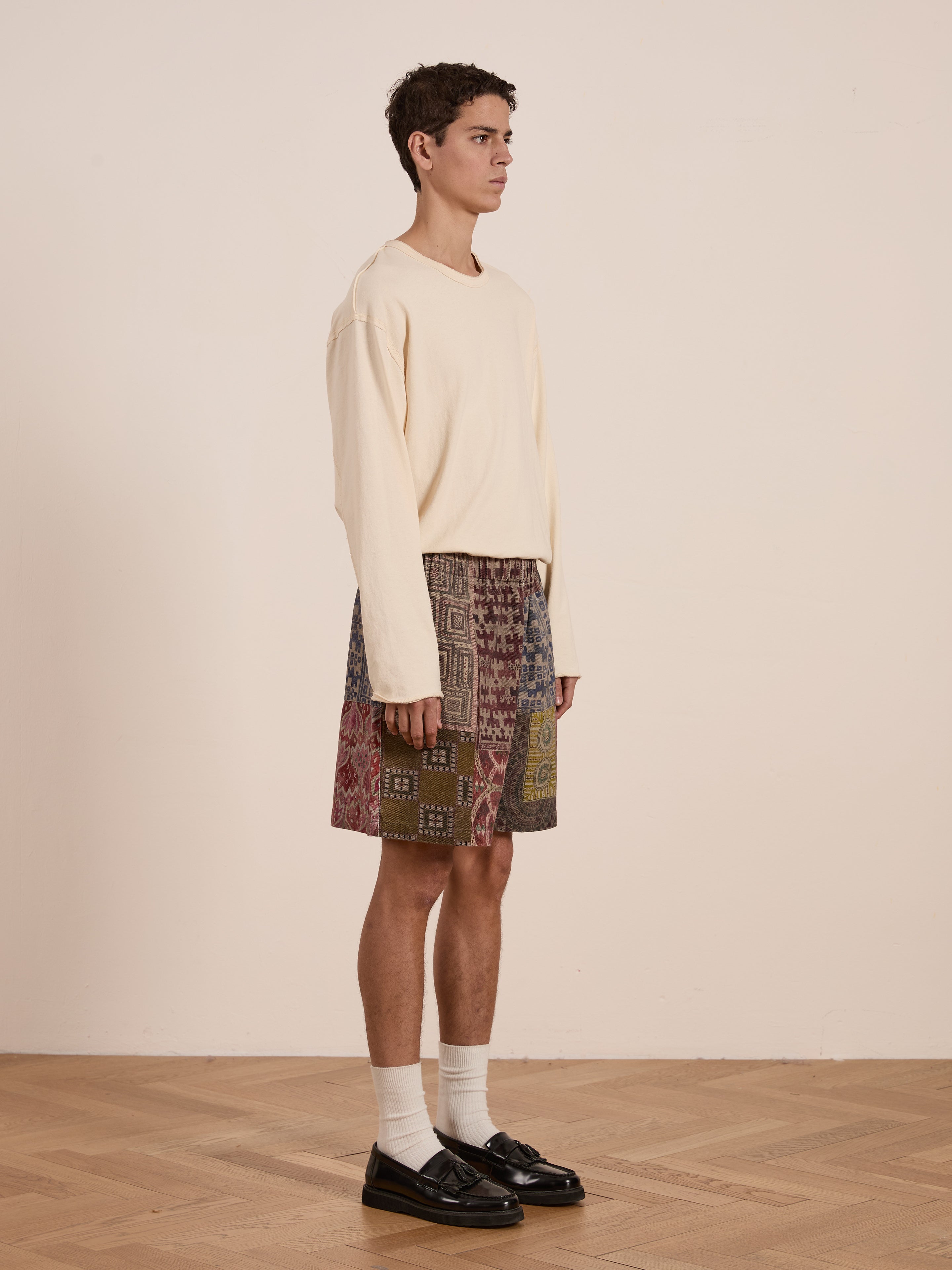 A person stands on a wooden floor against a plain white wall, wearing a beige long-sleeve shirt paired with FOUND's Watercolor Mosaic Work Shorts. Complementing the look are white socks and black loafers, embodying a unisex relaxed fit with artistic flair.