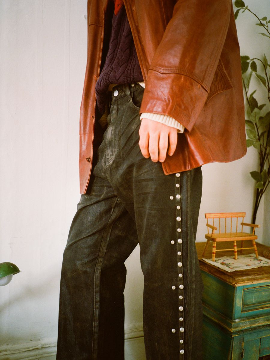 A person stands beside a small turquoise cabinet and a plant, wearing FOUND's Waxed Studded Jeans and a leather jacket, showcasing an appreciation for vintage craftsmanship.