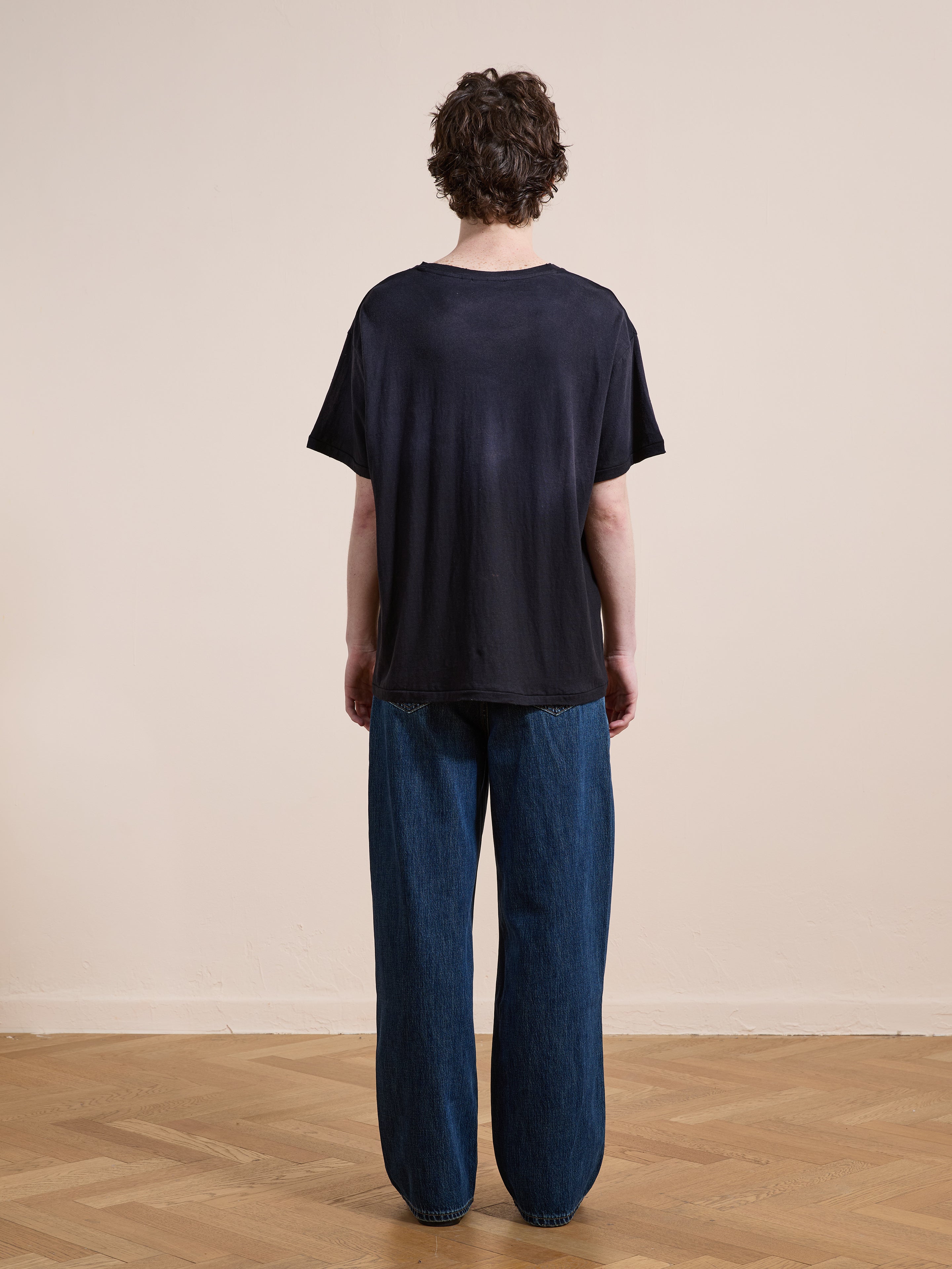 A person with short curly hair faces away, wearing FOUND's Weathered Pocket Tee in a unisex relaxed fit and wide-leg blue jeans on a wooden floor against a plain beige wall.