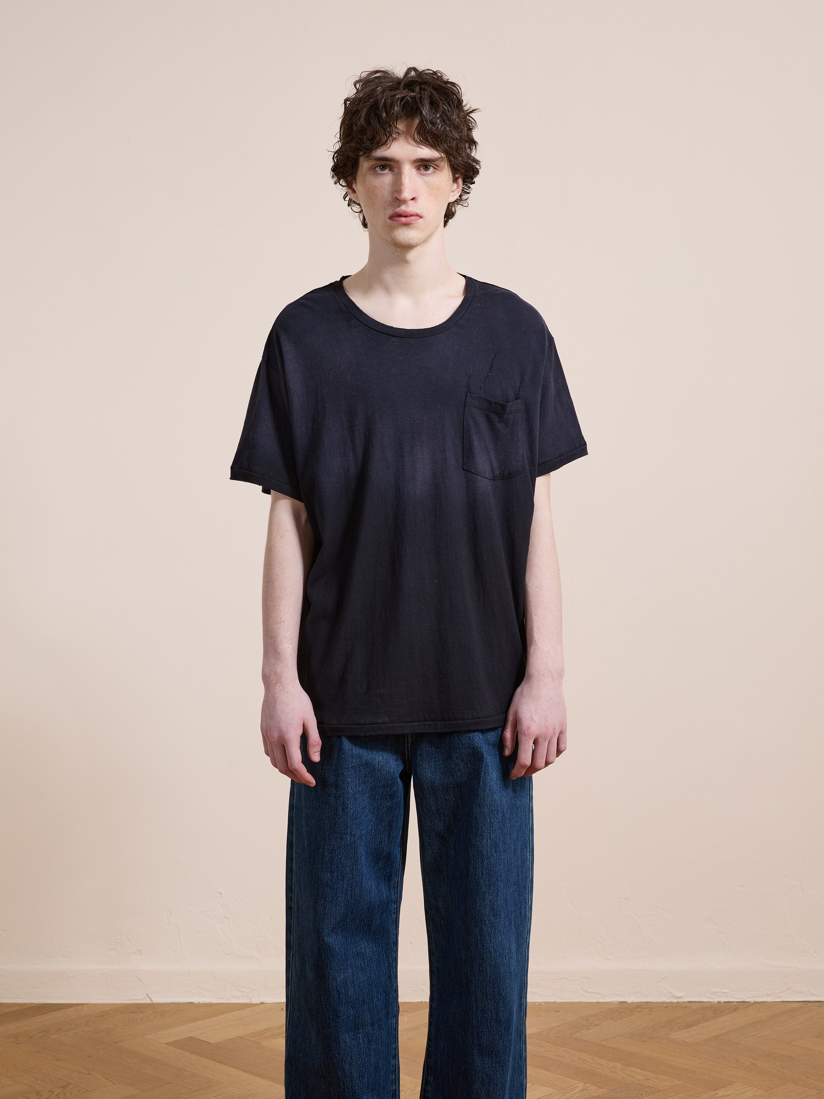 A person with curly hair stands indoors against a plain beige wall, wearing the FOUND Weathered Pocket Tee in black and blue jeans, looking directly at the camera.