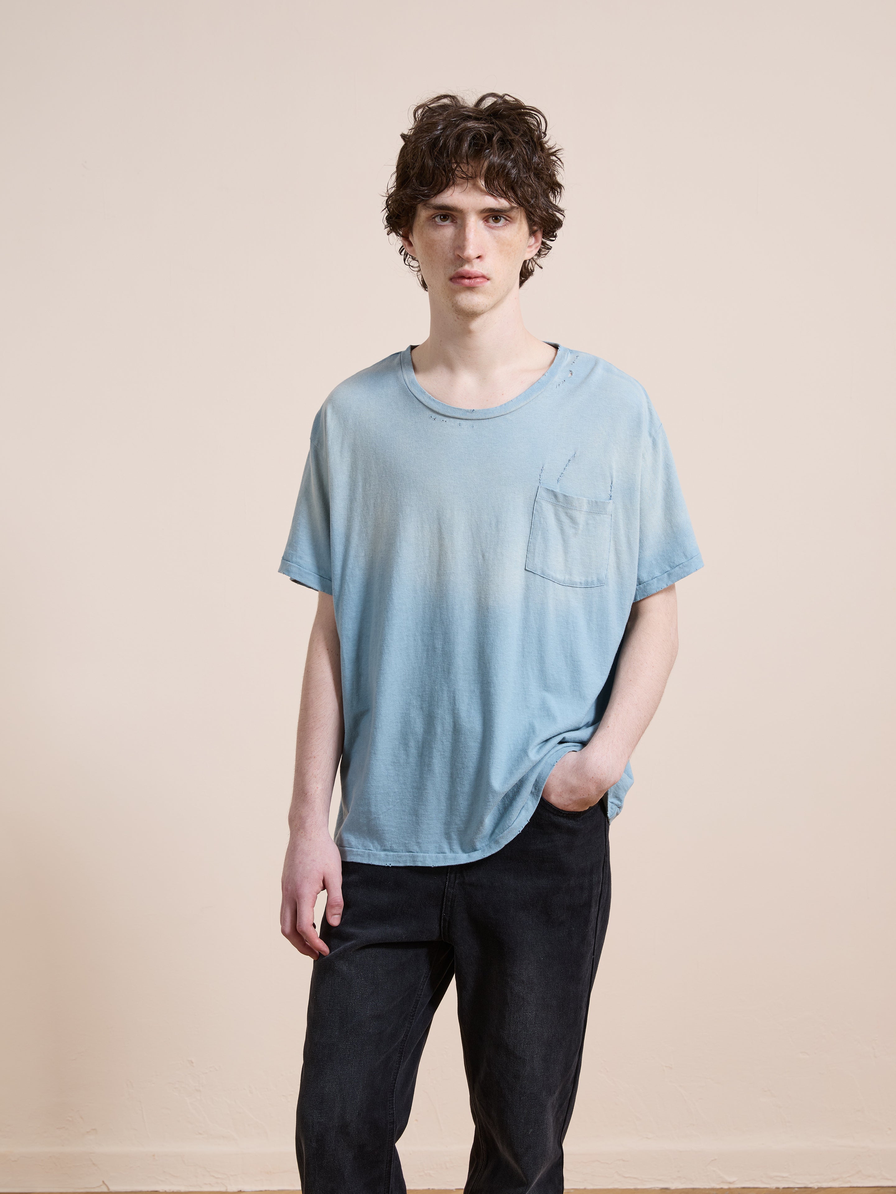 A person with wavy brown hair wears the FOUND Weathered Pocket Tee in a relaxed unisex fit and black pants, standing against a plain beige background.