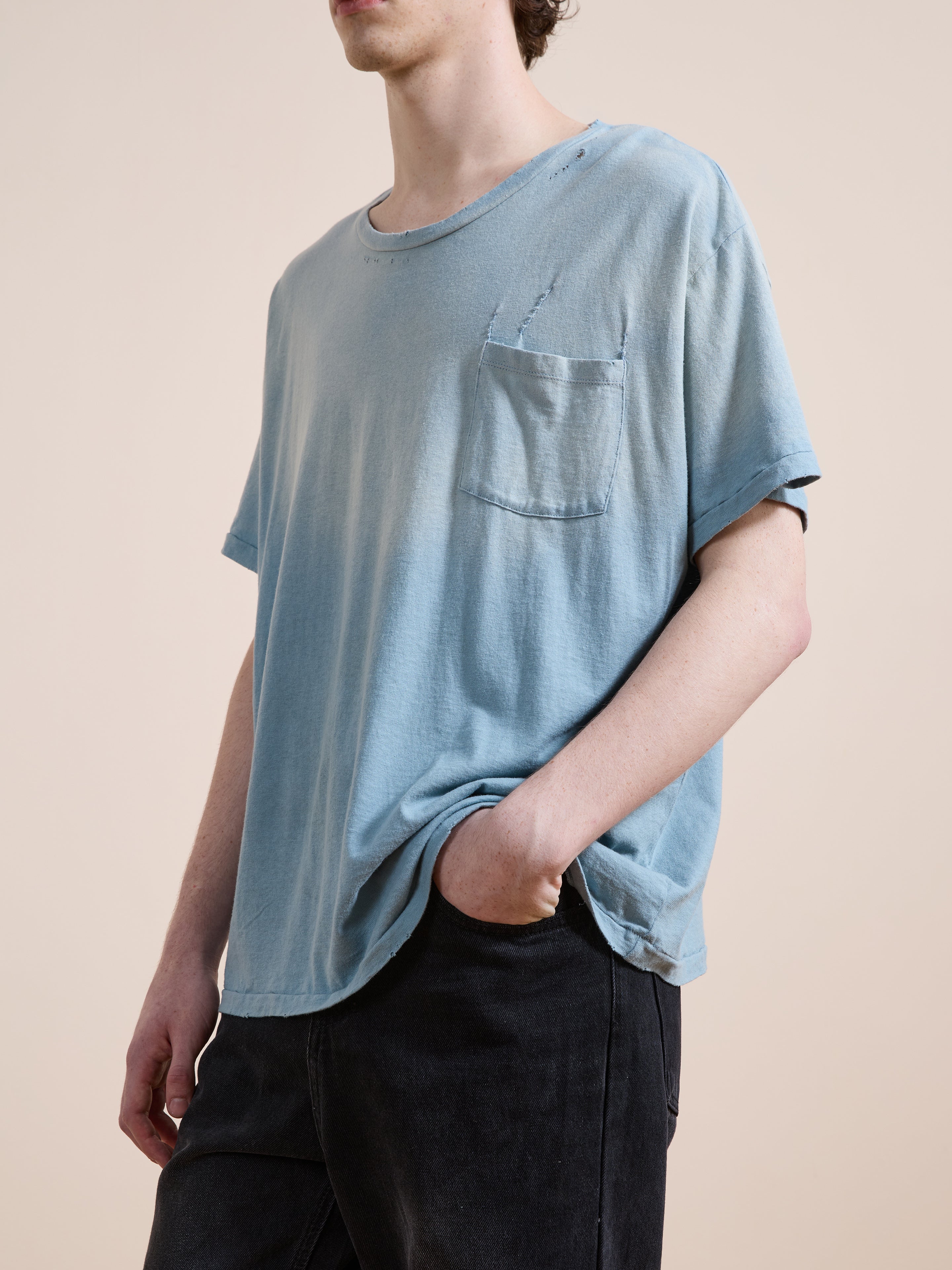 Someone wears the FOUND Weathered Pocket Tee—a loose, light blue vintage-style cotton shirt with chest pocket and visible distressing—paired with black pants, standing against a plain background.