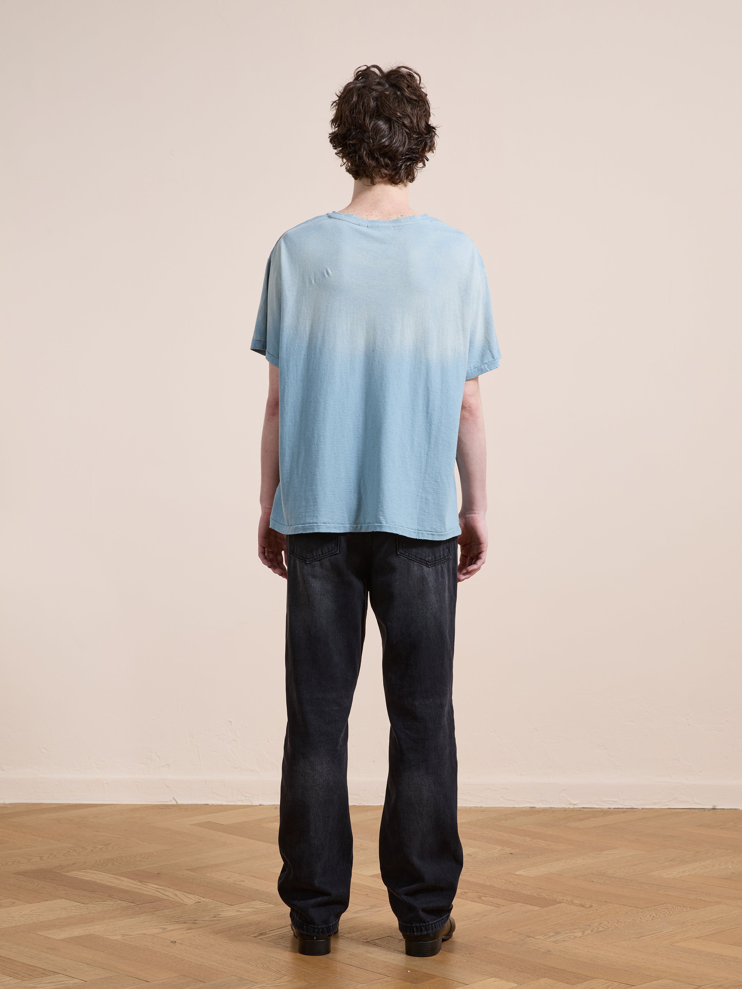 A person stands indoors facing away, wearing the FOUND Weathered Pocket Tee—a light blue unisex relaxed-fit shirt—paired with dark jeans on a wooden floor against a plain beige wall.