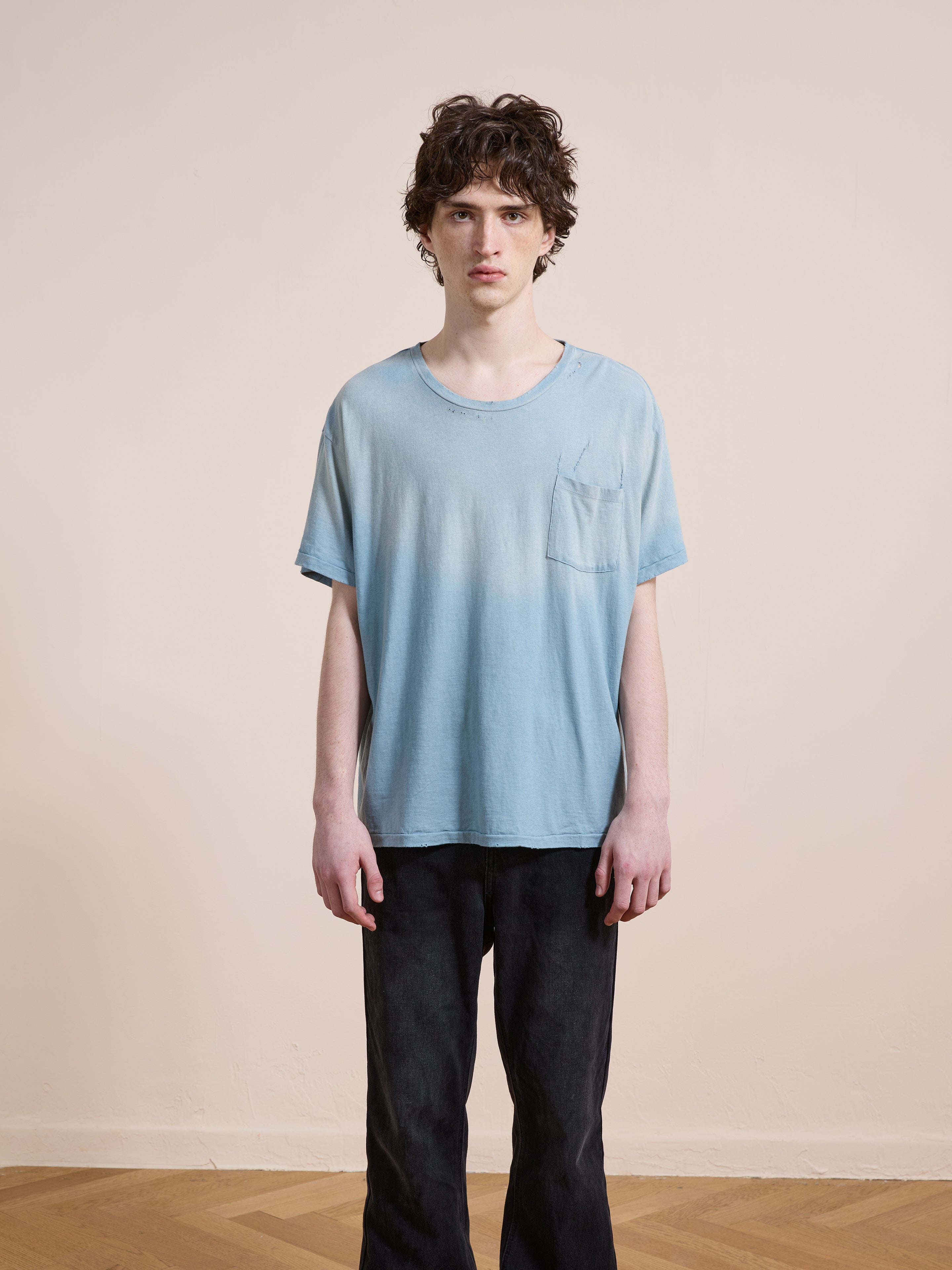 A young person with curly brown hair stands indoors, facing the camera in FOUND’s Weathered Pocket Tee—a relaxed fit, light blue vintage-style shirt—paired with black pants and a neutral expression.