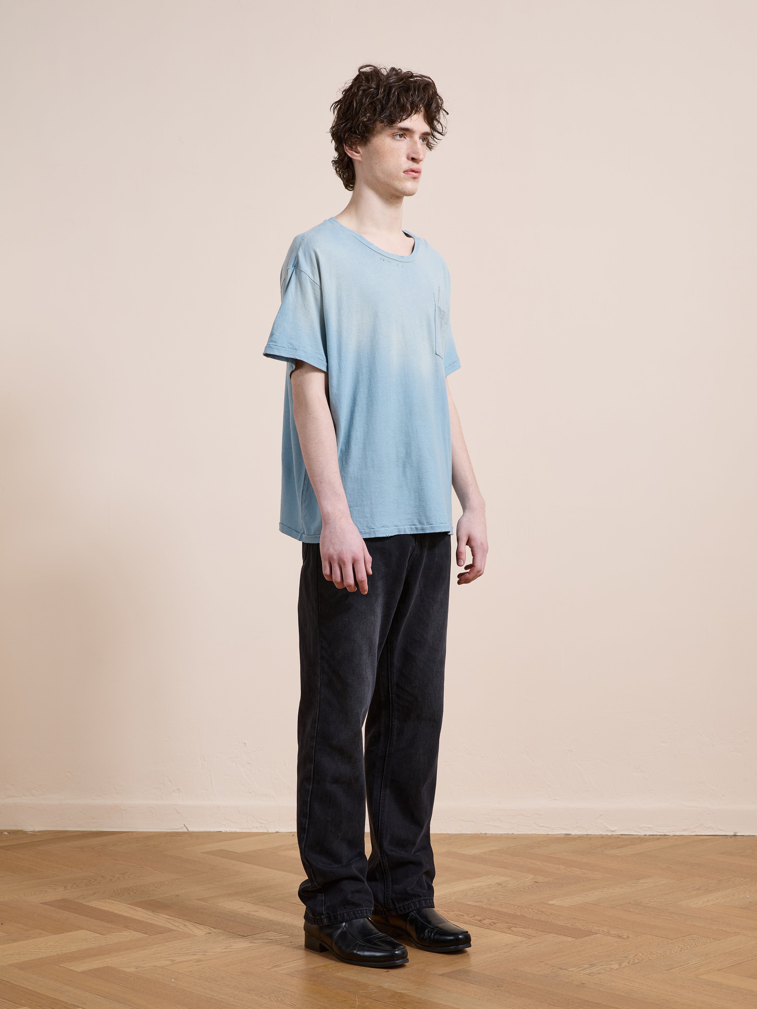 A person stands indoors on a wooden floor, wearing the FOUND Weathered Pocket Tee in light blue with dark pants and black shoes, facing slightly to the right against a plain beige wall.