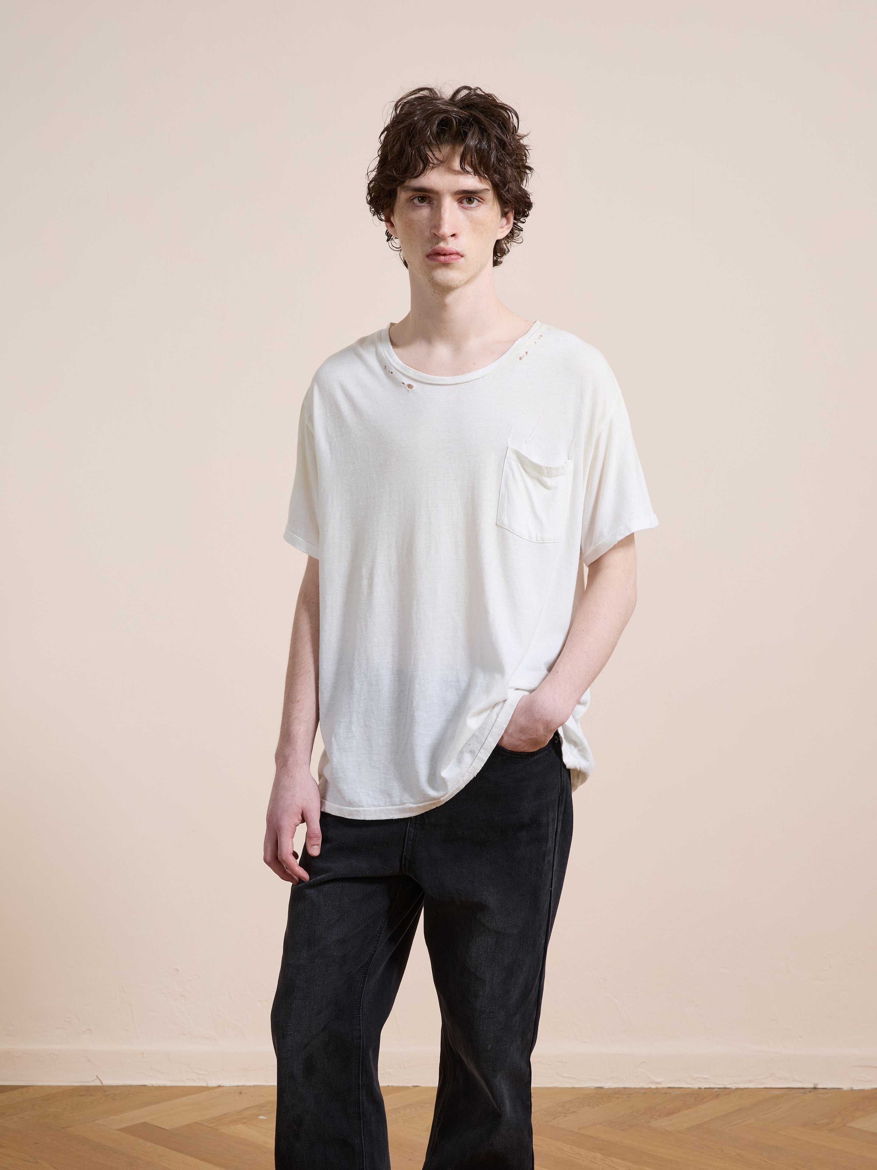 Indoors against a plain wall, a person wears FOUND’s Weathered Pocket Tee—crafted in soft cotton with vintage details and small holes—paired with dark pants and one hand casually slipped into a pocket.