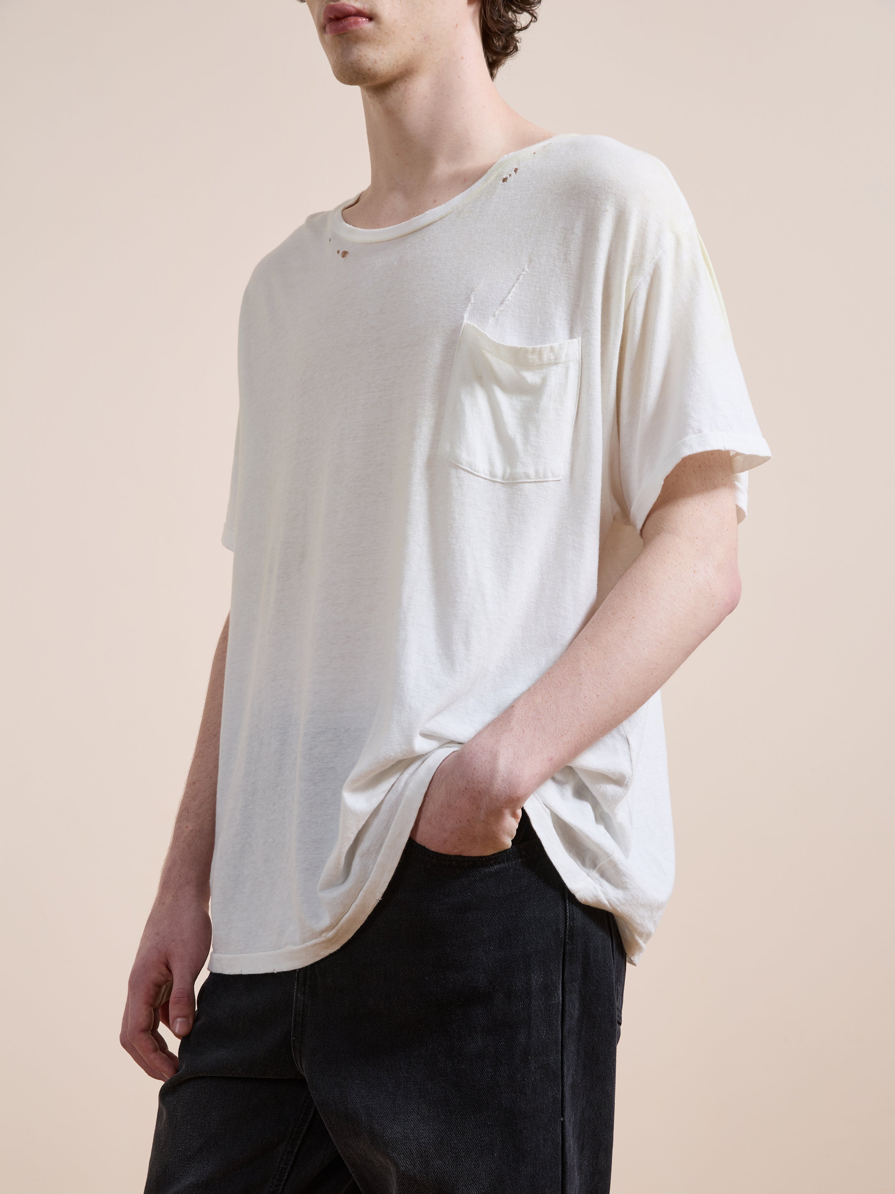 A person wearing the FOUND Weathered Pocket Tee—a unisex, relaxed fit white vintage T-shirt made from soft cotton with a chest pocket and subtle distressing—paired with black pants and standing against a plain background.